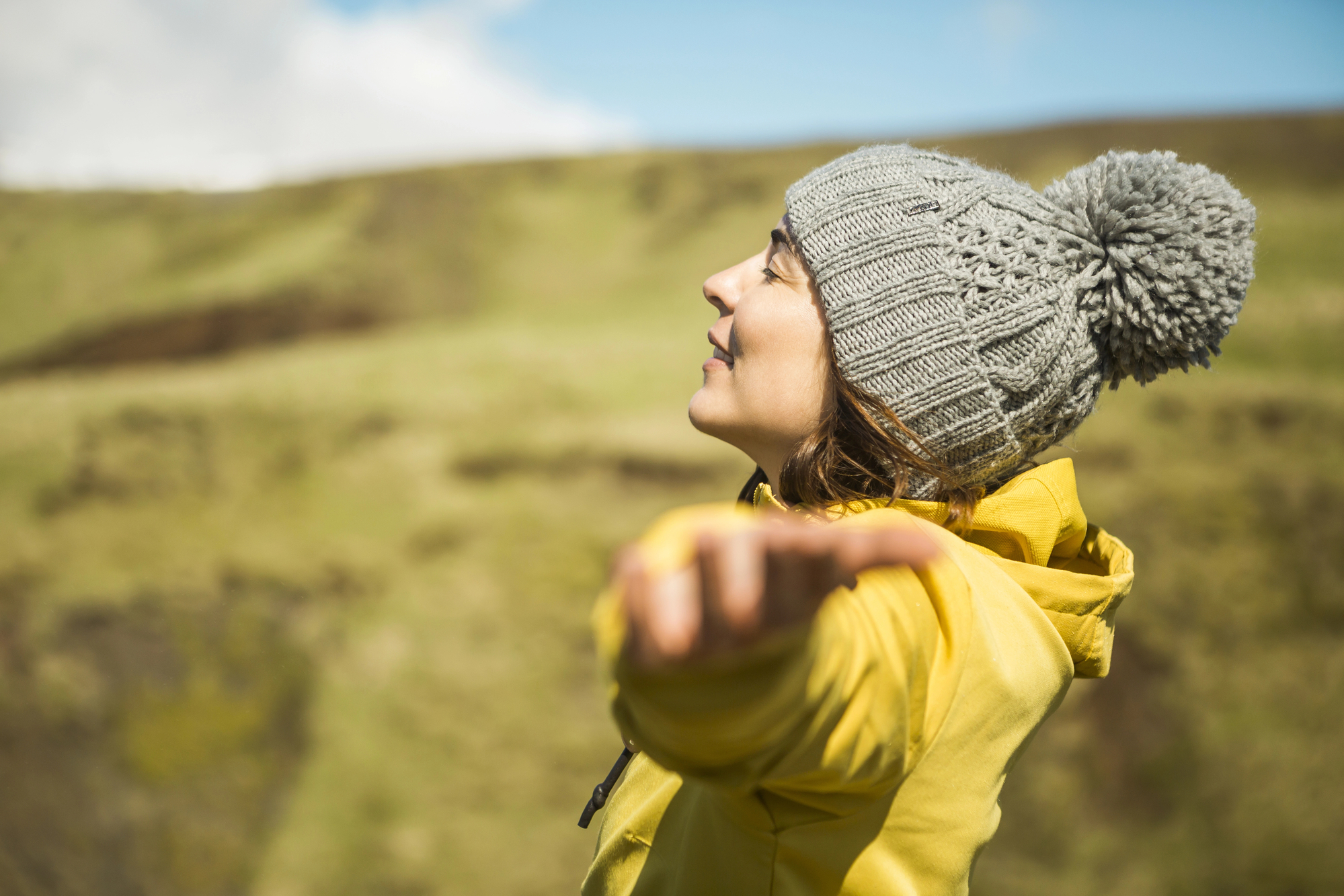 A person wearing a gray knit hat with a pom-pom and a yellow jacket stands outdoors with their eyes closed and arms outstretched, enjoying the sun. They are in a grassy landscape with a blurred background of hills and a partly cloudy sky.