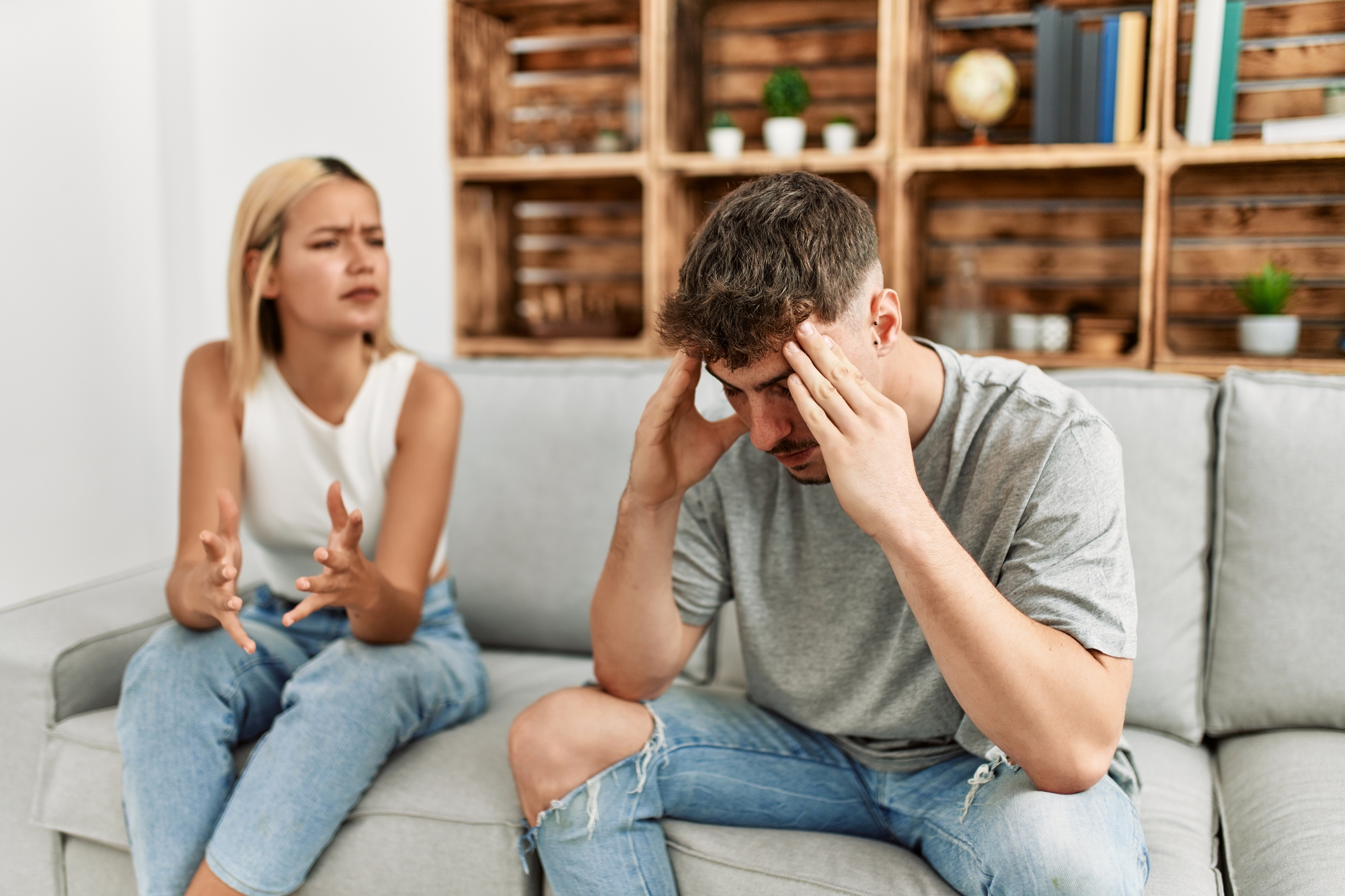 A woman and a man sitting on a gray couch appear to be in a heated discussion. The woman, with a frustrated expression, gestures with her hands, while the man holds his head in his hands, looking distressed. A wooden shelf with books and plants is in the background.