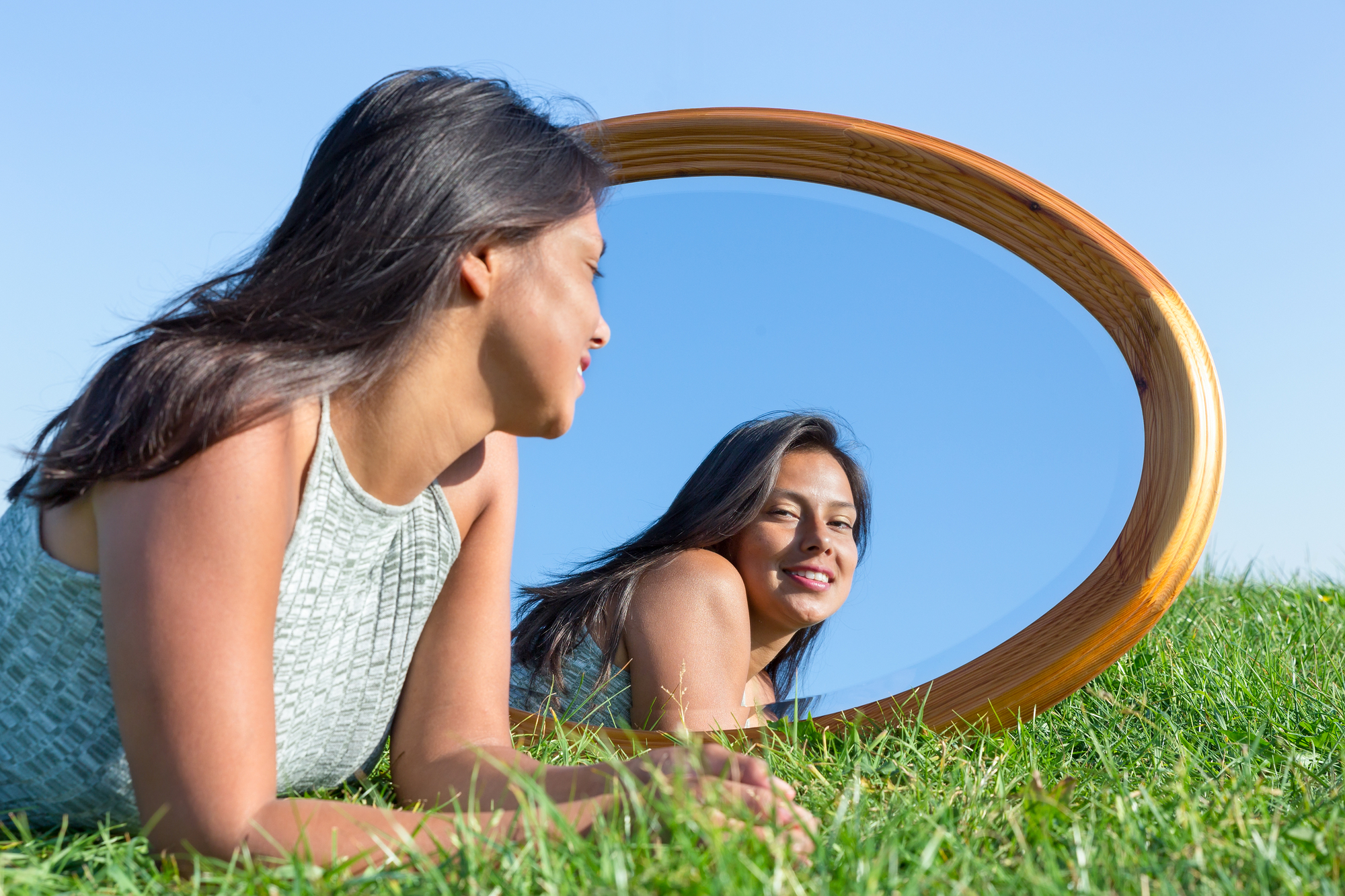 A woman with long dark hair is laying on green grass, facing a large oval mirror that rests on the ground. She is wearing a light green sleeveless top and smiling at her reflection. The sky is bright blue and clear.