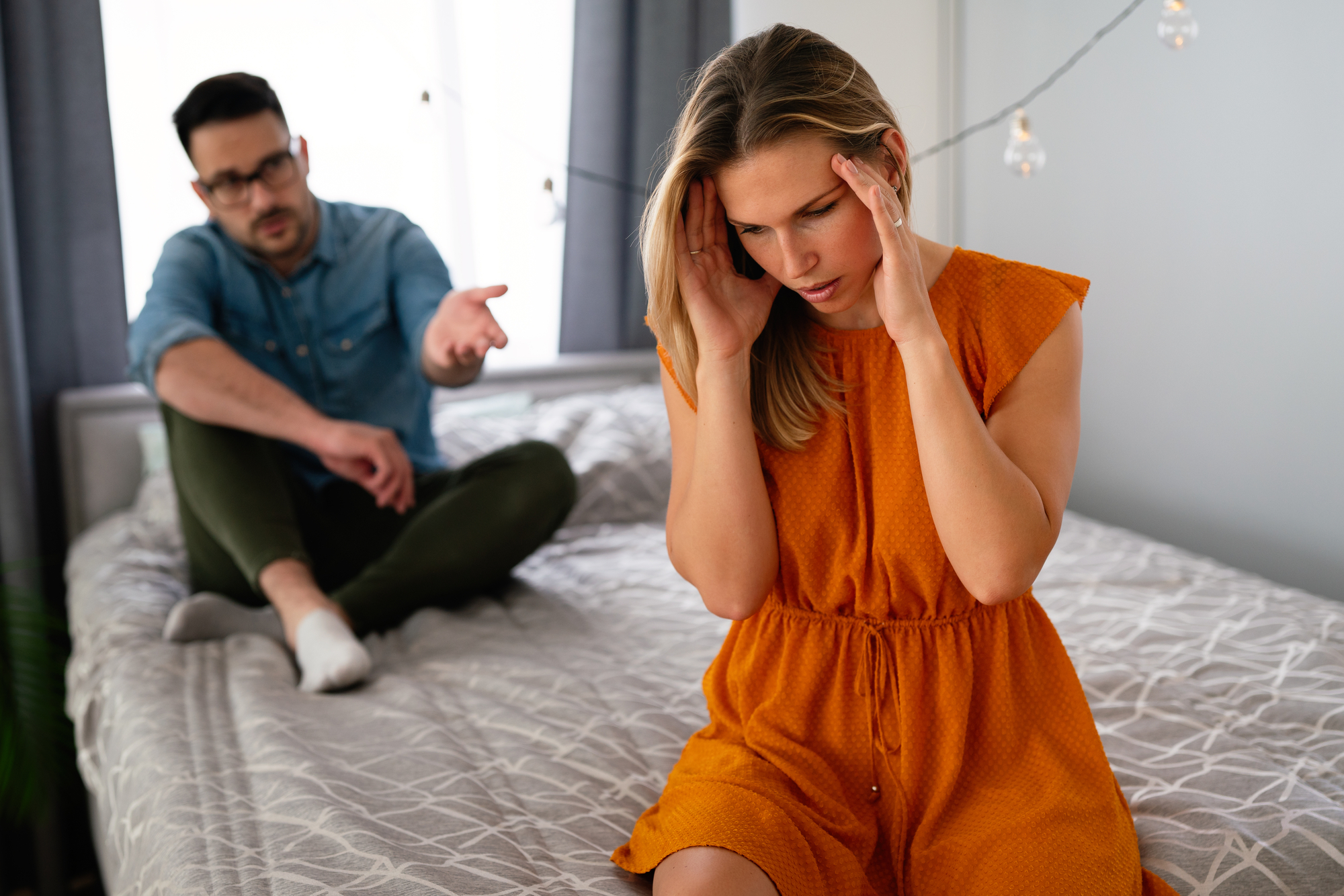 A woman in an orange dress sits on the edge of a bed, holding her head in her hands, appearing distressed. A man in glasses and casual clothes sits behind her on the bed, extending his arm as if explaining or trying to communicate with her.