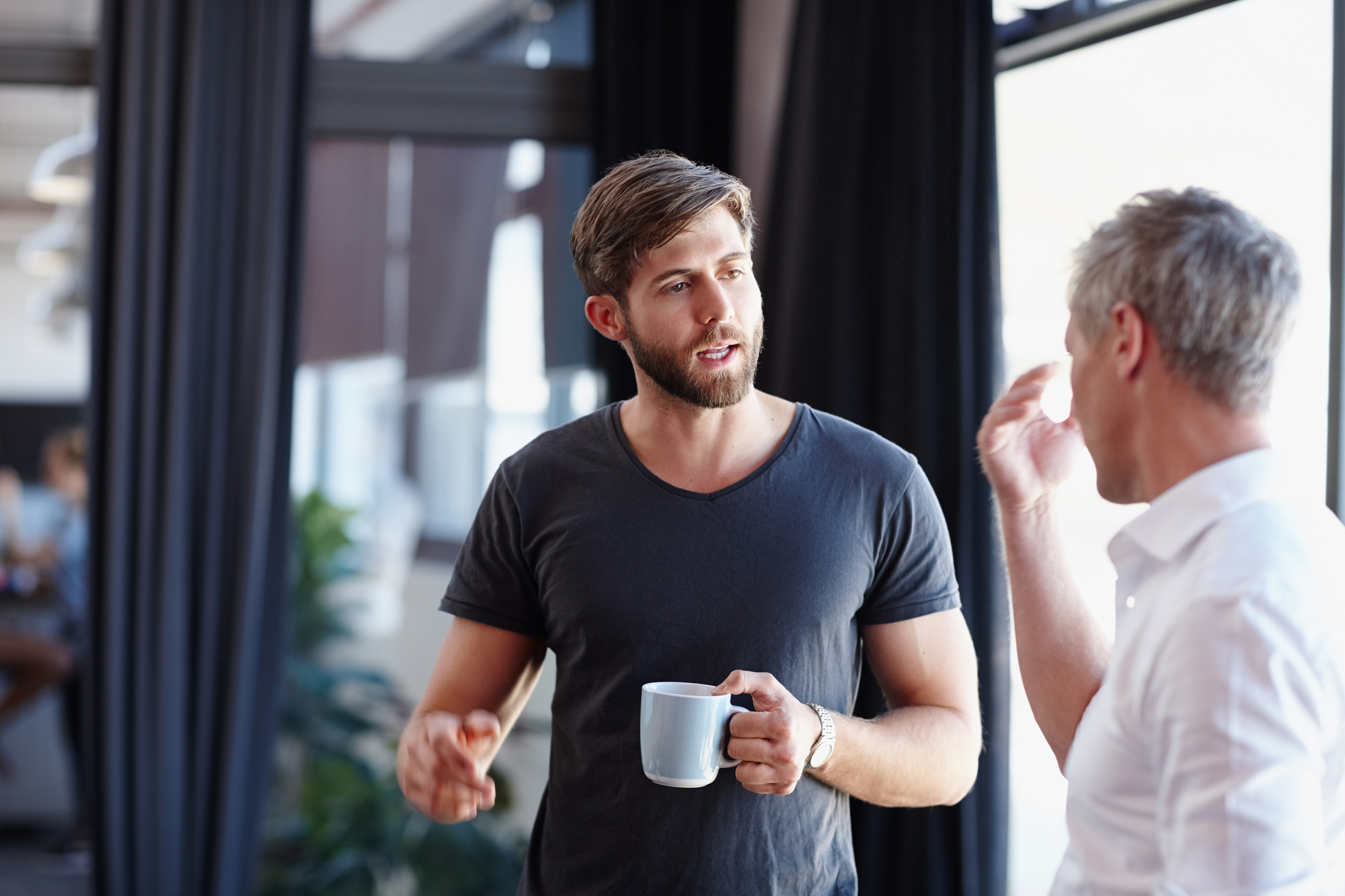 Two individuals are engaged in a conversation indoors. One person, holding a white mug, appears to be speaking, while the other individual is listening attentively. They are dressed casually, and the setting features large windows and modern decor.