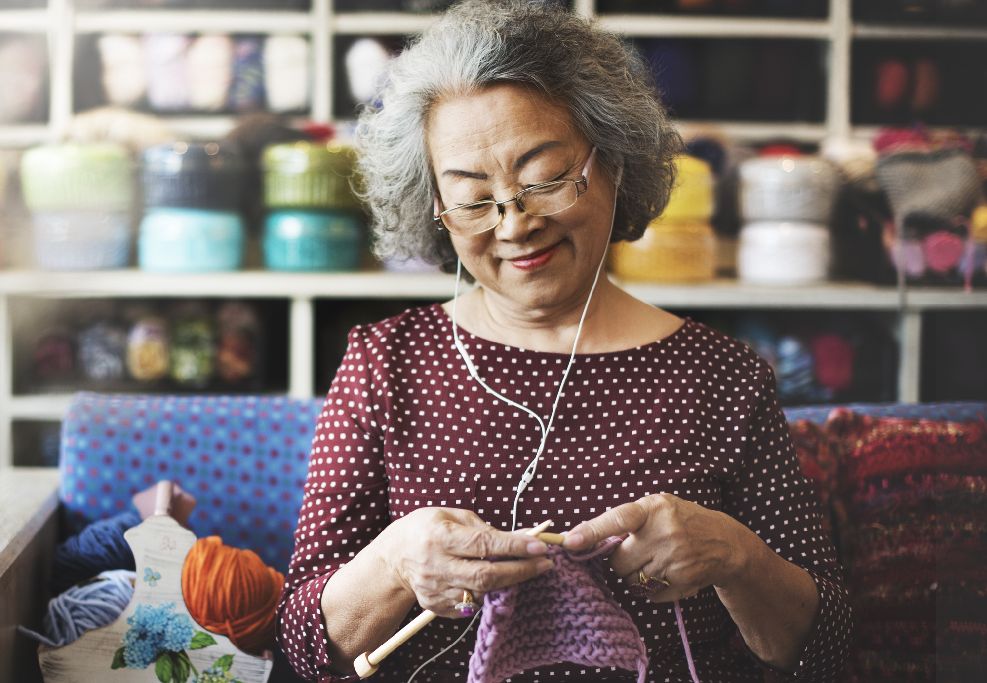 An elderly woman with gray hair and glasses, wearing a polka dot blouse, is knitting with purple yarn. She is listening to something through earphones. Behind her are shelves filled with colorful yarn. A basket of yarn is on the table next to her.