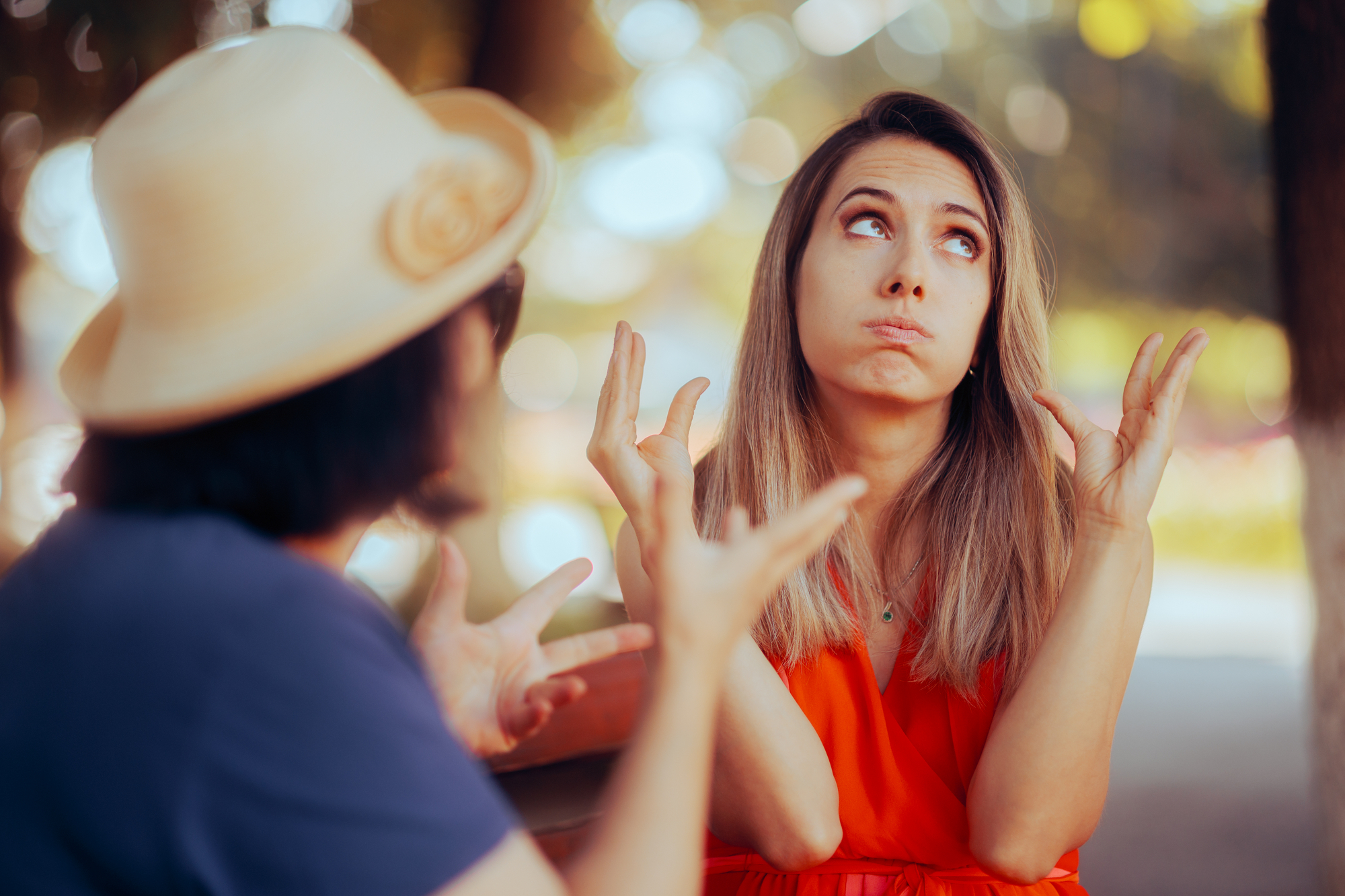 Two women engage in an animated conversation outdoors. One woman, wearing a hat, gestures expressively. The other woman, in a red top, looks up with an exasperated expression, her cheeks puffed out and hands raised in a frustrated gesture.