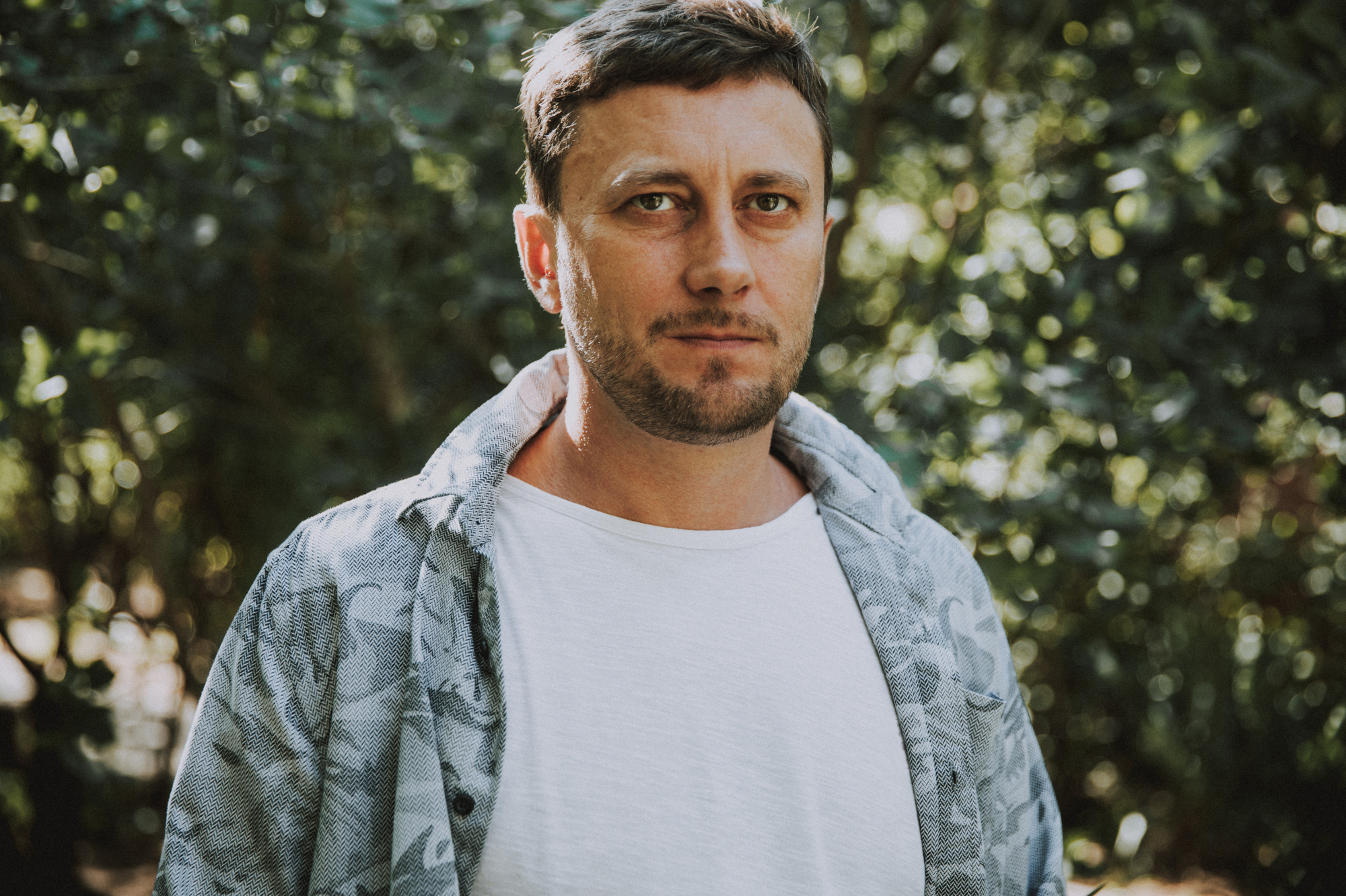 A man with short brown hair and light facial hair stands outdoors in front of green foliage. He is wearing a white t-shirt under a patterned denim jacket and gazes directly at the camera with a neutral expression. The sunlight filters through the trees, creating a natural, serene background.