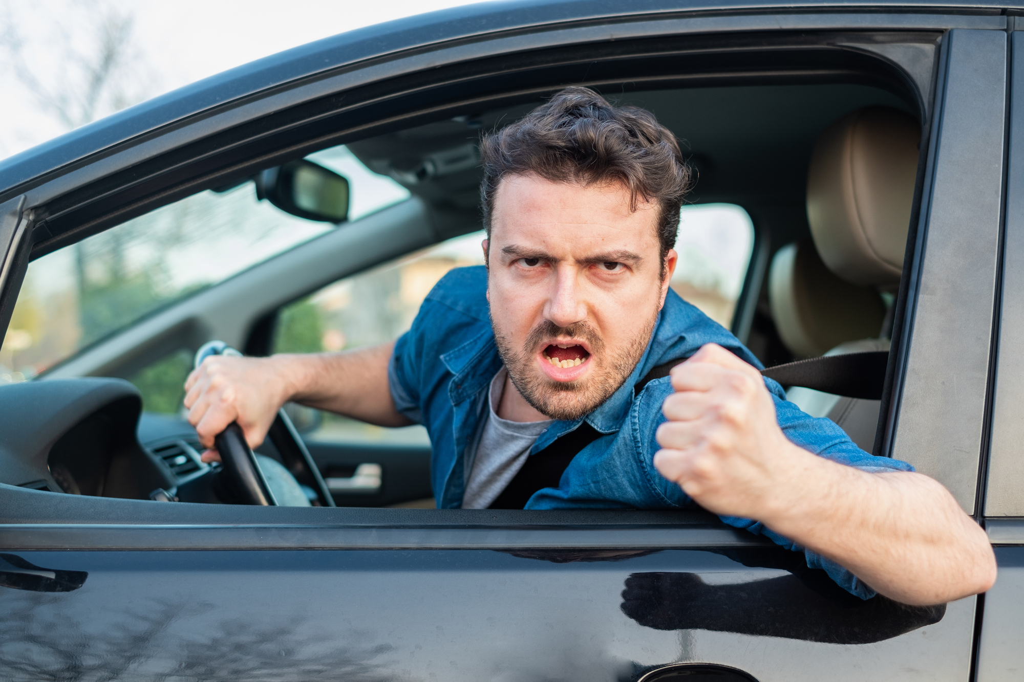 A man in a blue shirt angrily leans out of the driver's seat window of a car with a frustrated expression, shaking his fist. The background is blurred, indicating an outdoor setting.