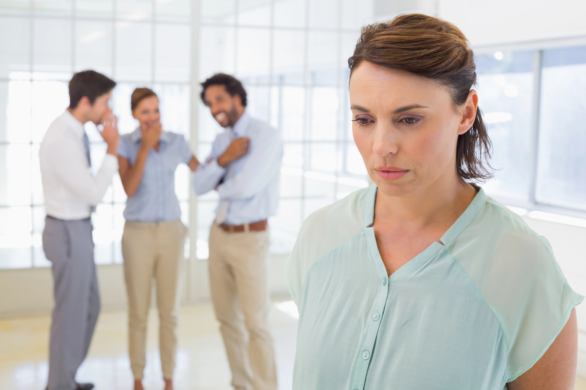 A woman stands in the foreground looking sad and concerned while three colleagues in the background whisper and laugh together in an office setting with glass walls.