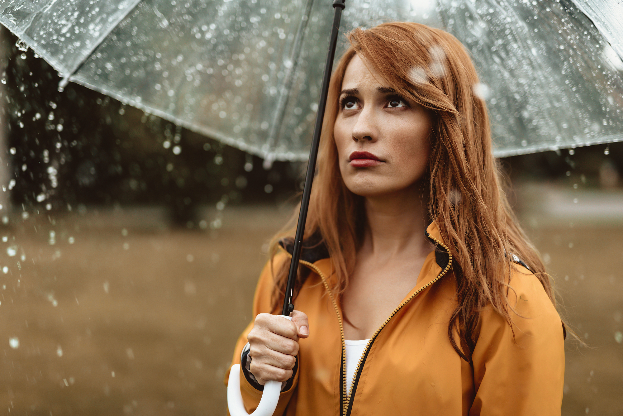 A woman with long brown hair stands outdoors under a clear umbrella, wearing a yellow raincoat. She looks upwards with a slightly displeased expression as raindrops fall around her. The background is blurred, showing a natural, possibly park-like setting.