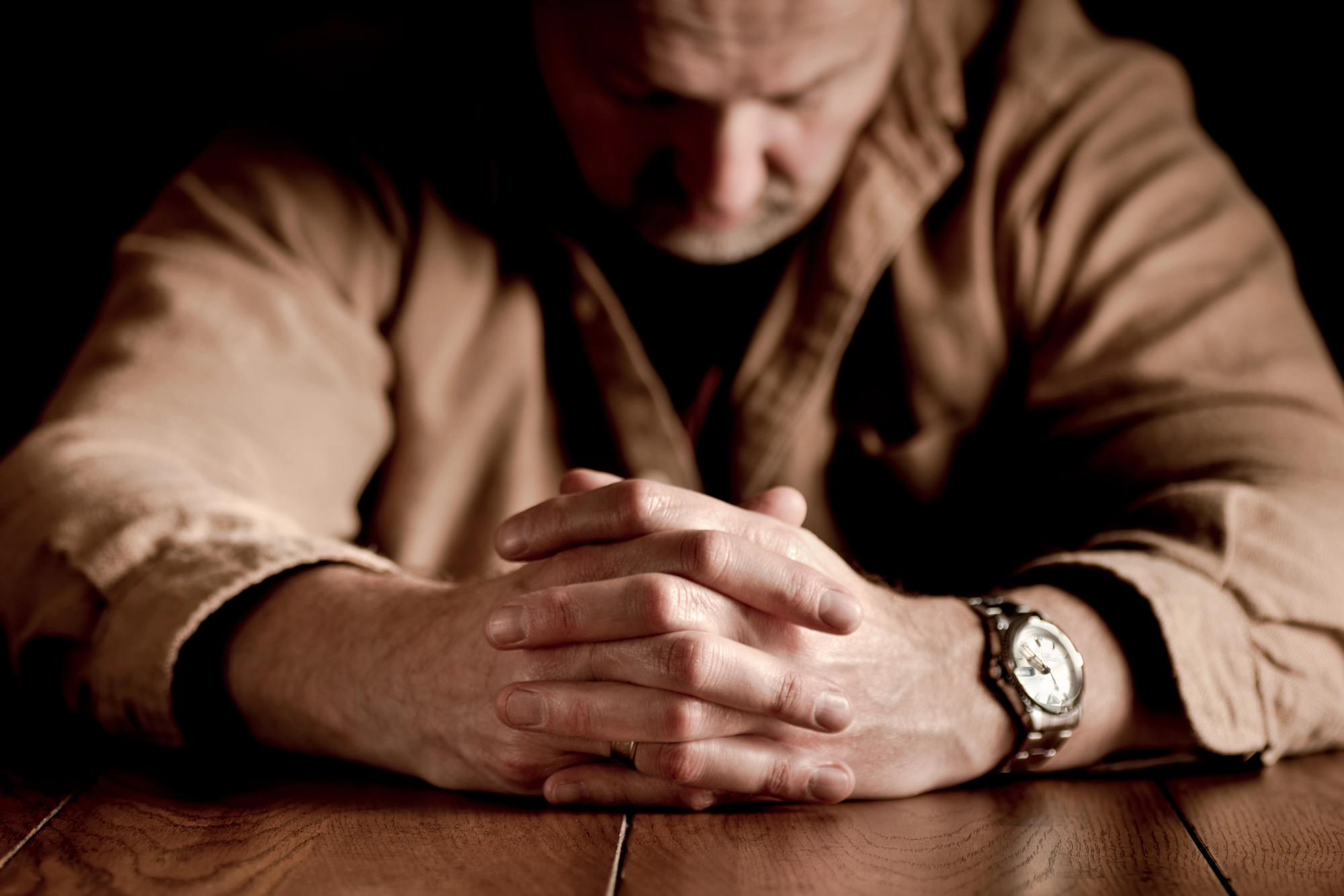 A man wearing a brown shirt sits at a wooden table with his hands clasped together. His head is bowed, partially obscured, and he appears to be deep in thought or prayer. A watch is visible on his left wrist. The lighting is dim, creating a contemplative atmosphere.
