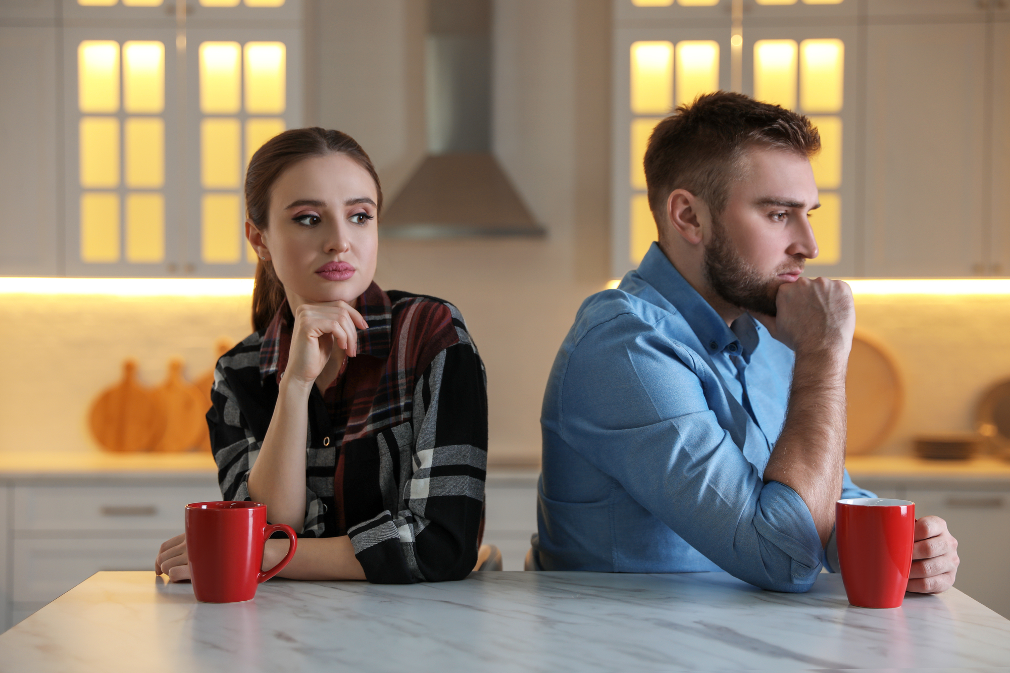 A woman and a man sit back-to-back at a kitchen counter, appearing upset with each other. Both have thoughtful expressions and rest their chins on their hands. Each has a red mug in front of them, and the background shows a modern kitchen with warm lighting.