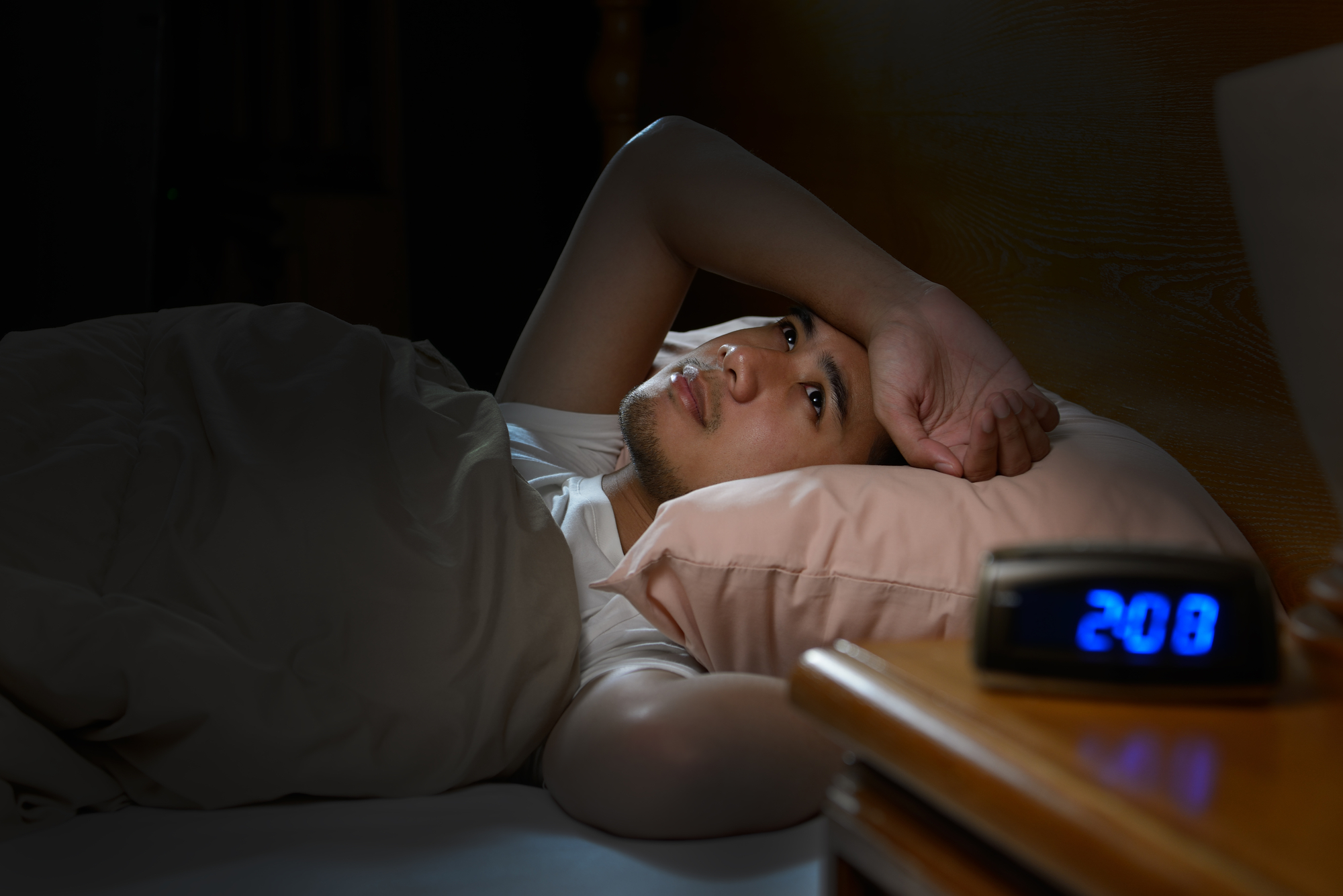 A person lies in bed, appearing awake and staring at the ceiling. They are partially covered with a blanket and rest their head on a pillow. Beside them, an alarm clock on the nightstand displays the time as 2:08. The scene is dimly lit.