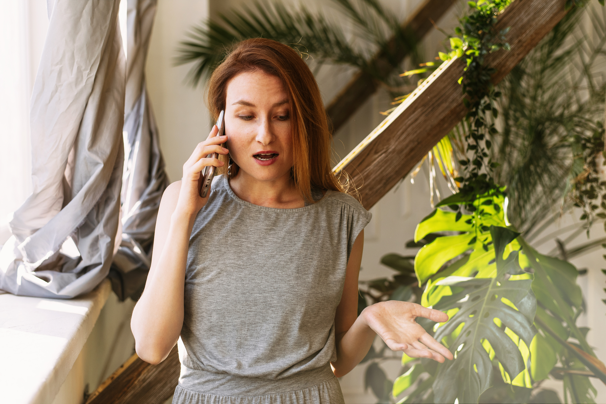 A woman with long red hair, wearing a grey dress, is indoors near a window with plants in the background. She holds a phone to her ear, looking surprised and gesturing with her other hand. Sunlight filters through the window and illuminates the scene.