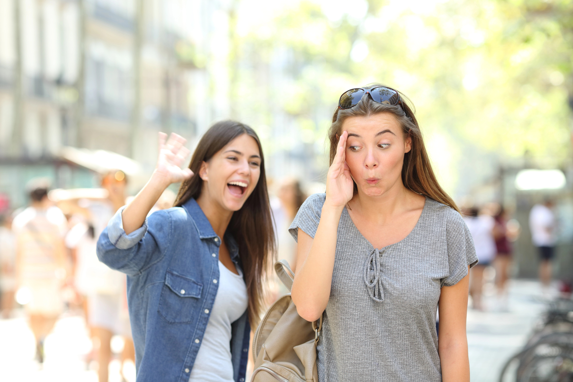 Two women are outdoors on a sunny day. The woman on the left is smiling and waving while wearing a denim shirt. The woman on the right, wearing a gray t-shirt and sunglasses on her head, is making a surprised face and touching her temple. Blurred street scene behind them.