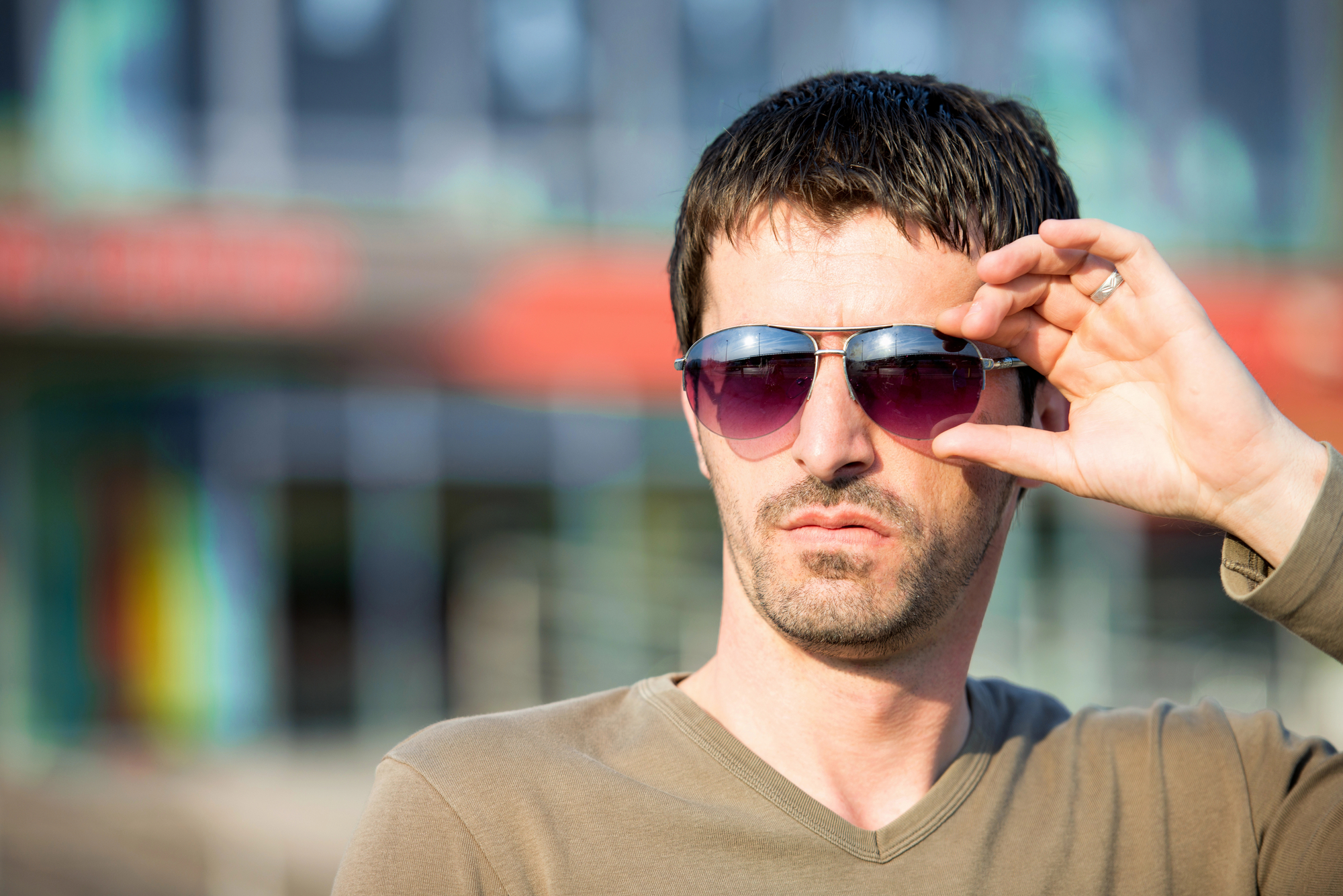 A man with short dark hair and a trimmed beard is standing outdoors, wearing a beige long-sleeve shirt and dark sunglasses. He is looking off into the distance while adjusting his sunglasses with one hand. The background is blurred, featuring some colorful elements.