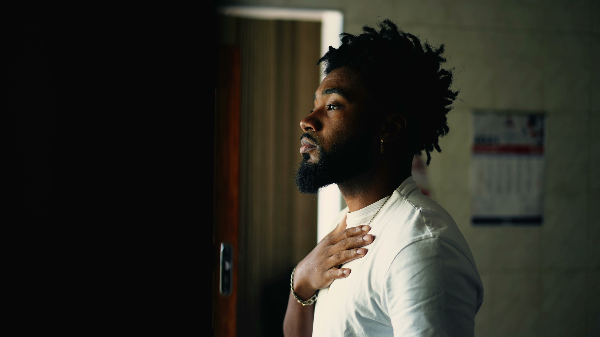 A man with a beard and curly hair stands indoors, lit from the side by natural light through a window. He wears a white shirt and a bracelet, and his hand rests on his chest. The background is dim, with a partially visible calendar on the wall.