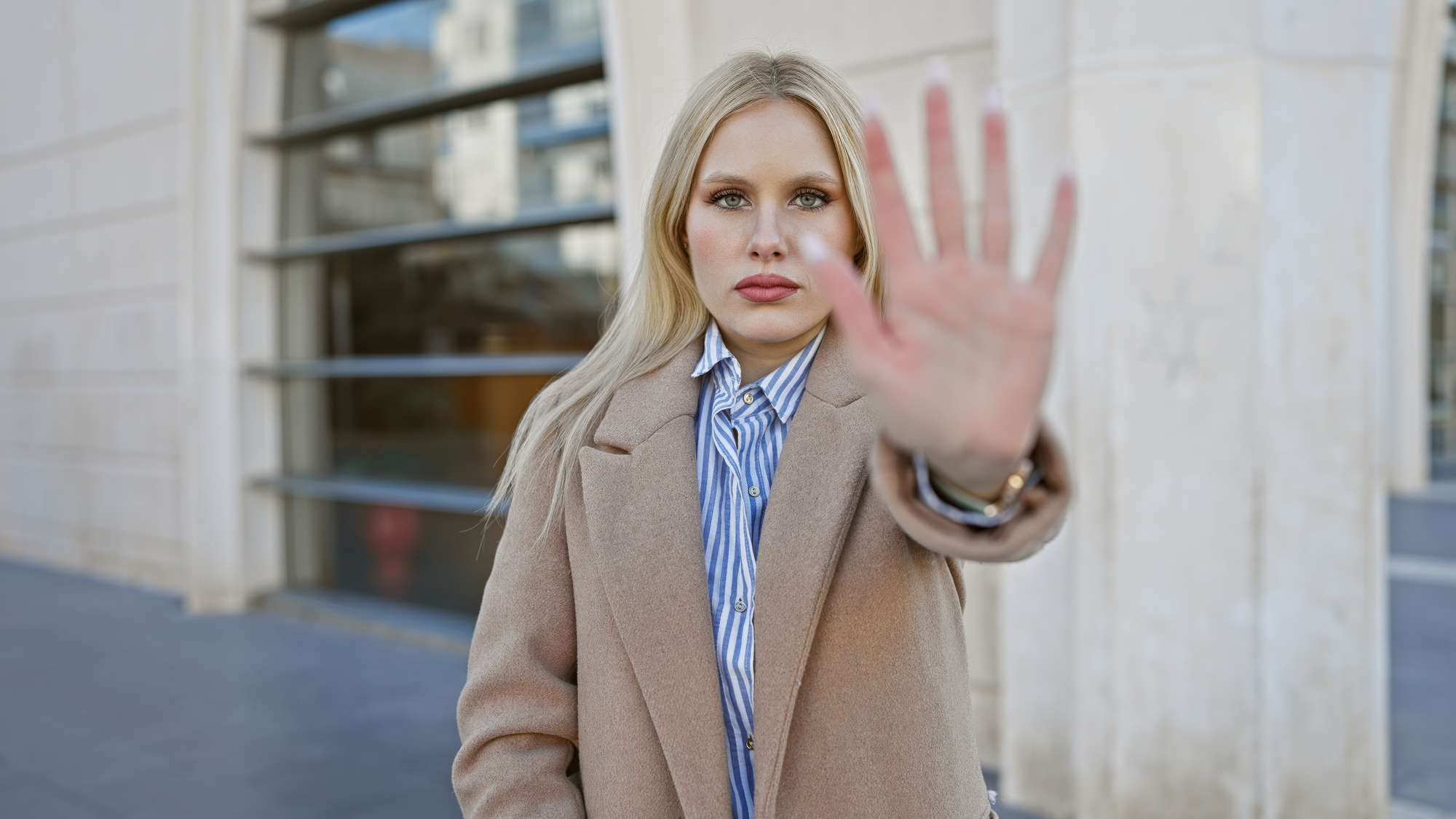 A woman with long blonde hair stands outside an urban building, looking directly at the camera. She is wearing a tan coat over a blue-and-white pinstriped shirt and holds her hand out in front of her, with her palm open and facing the camera.