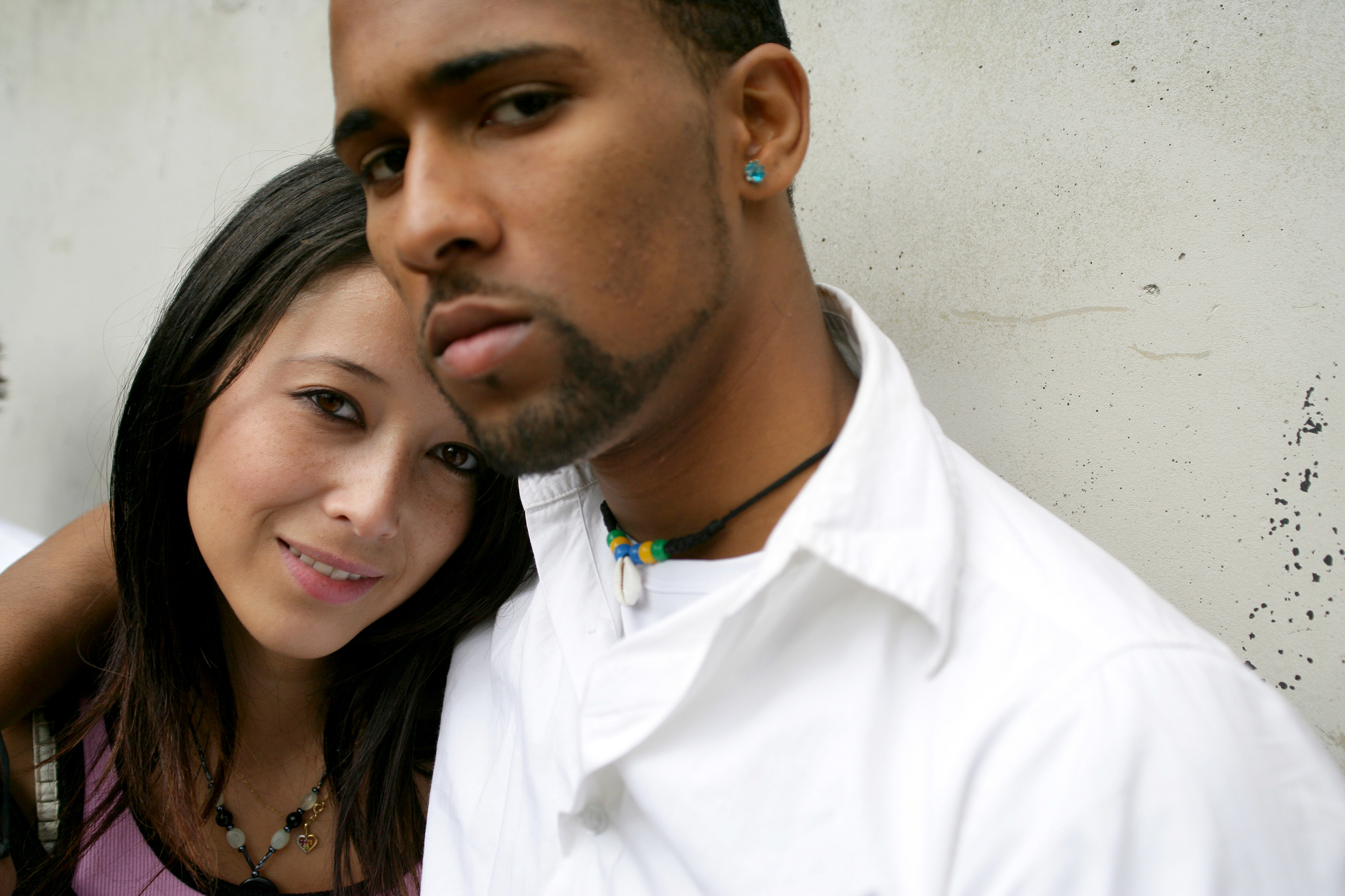 A close-up image of a young couple standing against a textured wall. The woman on the left has long dark hair, is smiling softly, and wears a pink top. The man on the right has short hair, a goatee, and wears a white shirt with a beaded necklace, looking directly at the camera.
