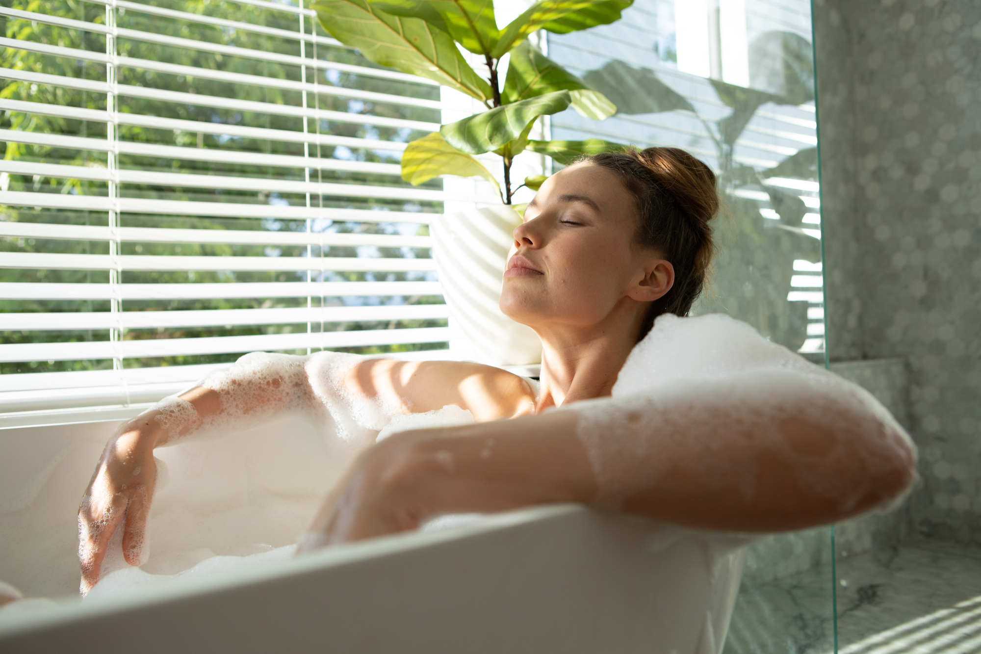 A person with eyes closed relaxes in a bubble bath, surrounded by sunlight streaming through white blinds. There is a green plant in a white pot next to the bathtub, creating a serene and peaceful atmosphere.