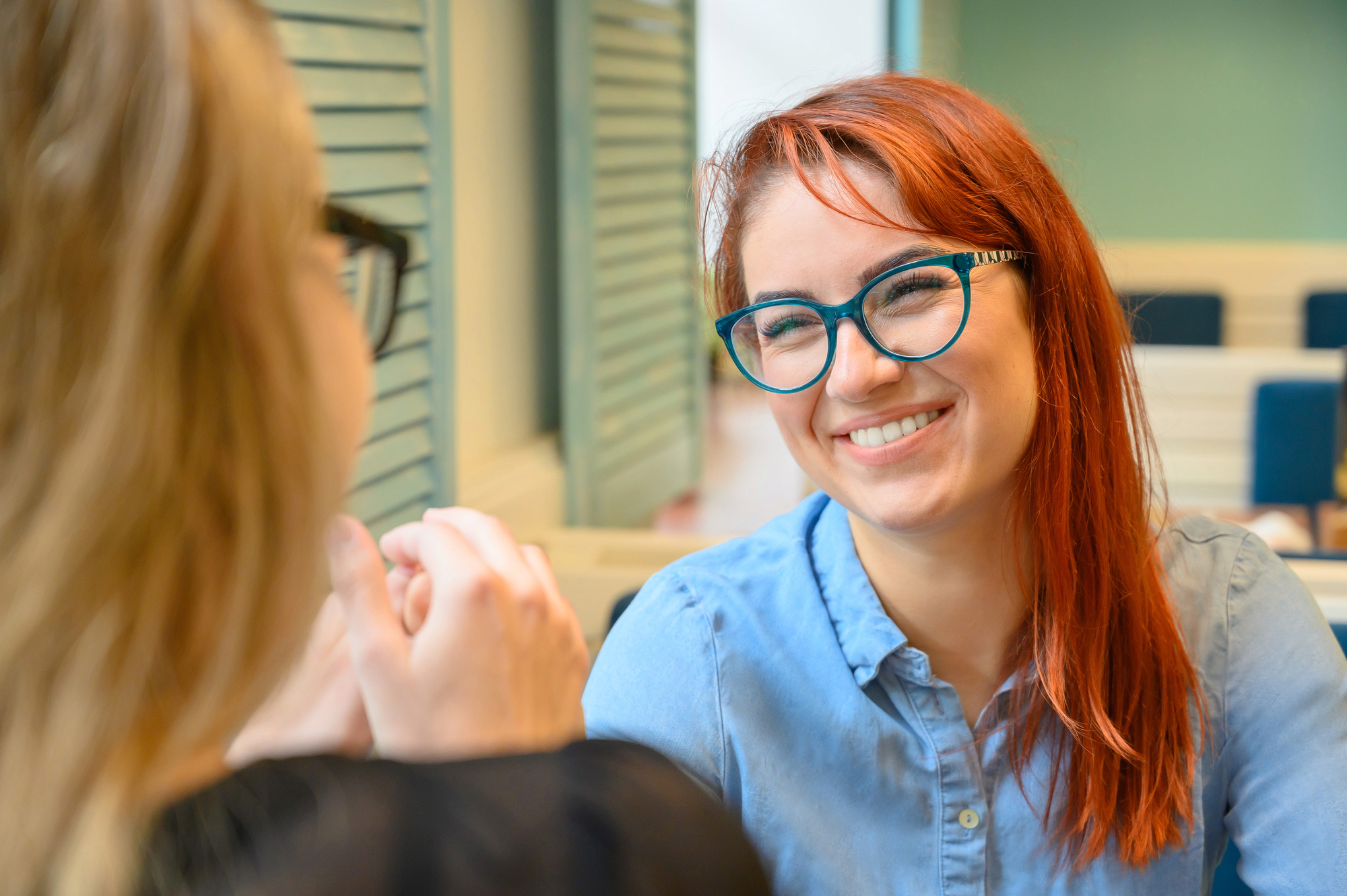 Two women sitting in a brightly lit room with blue accents, engaged in a cheerful conversation. The woman facing the camera has red hair, glasses, and is smiling, while the other woman, with blurred features, faces away from the camera.