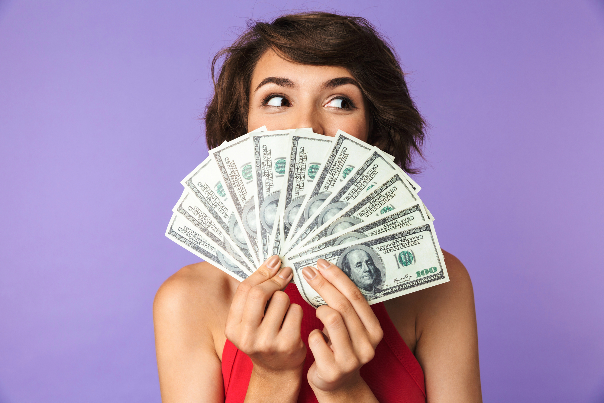 A person with short brown hair and a red sleeveless top holds up a fanned stack of hundred-dollar bills, partially covering their face, while looking to the side. The background is a solid purple color.