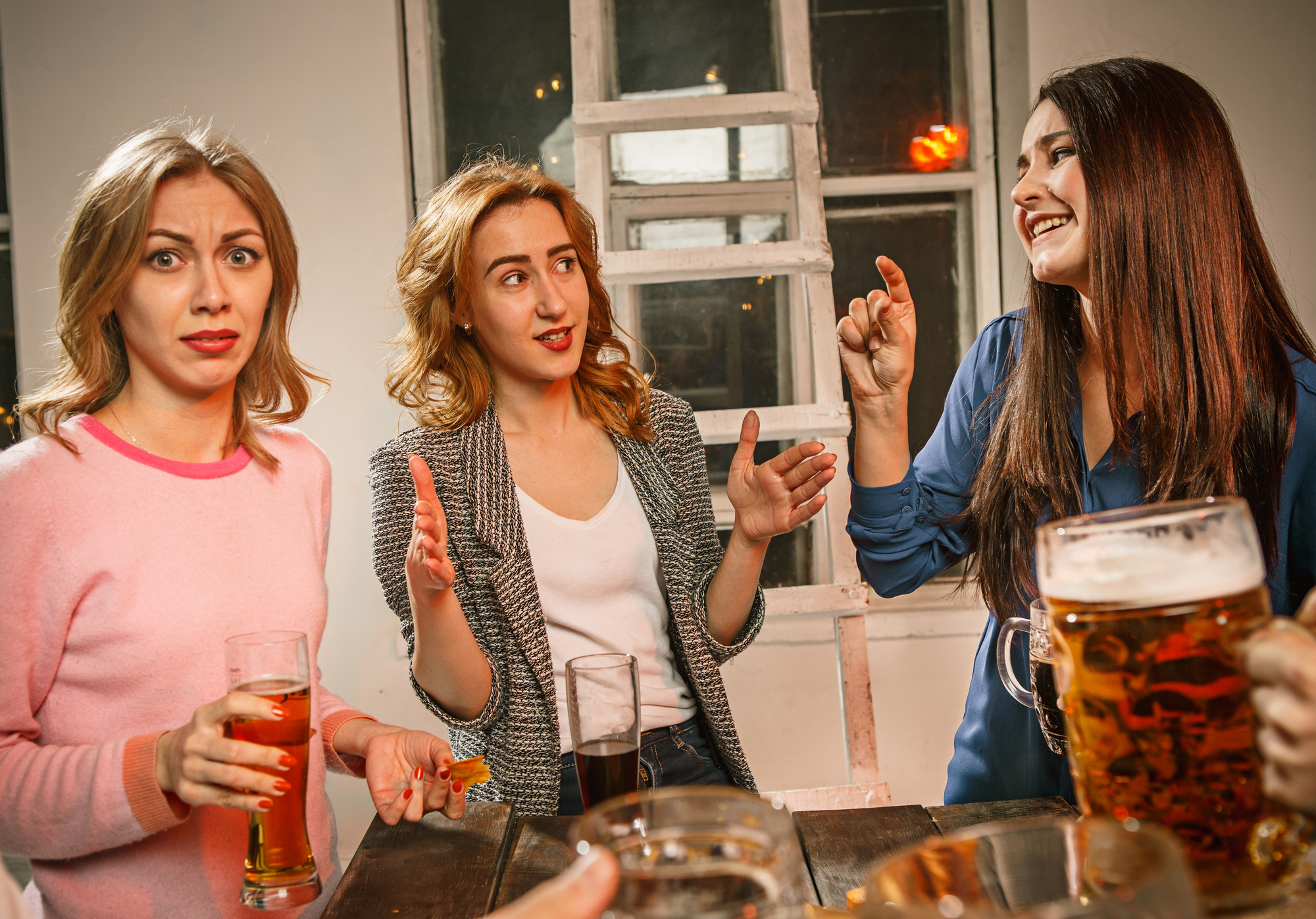 Three women stand around a table with drinks, engaged in animated conversation. One woman in pink looks surprised, the woman in the middle gestures with hands open, and the third woman in blue smiles while gesturing with her fingers. A hand holding a beer is in the foreground.