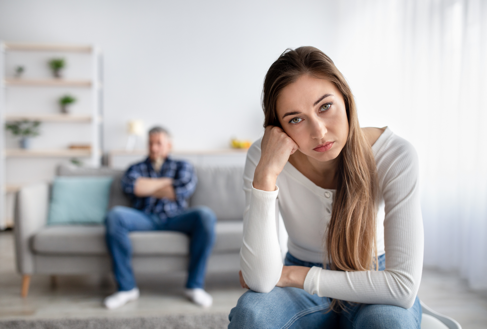 A woman with long brown hair, wearing a white long-sleeve shirt, rests her head on her hand and looks at the camera with a serious expression. In the background, a man sits on a couch with his arms crossed, looking away. The room has a cozy, neutral-toned decor.