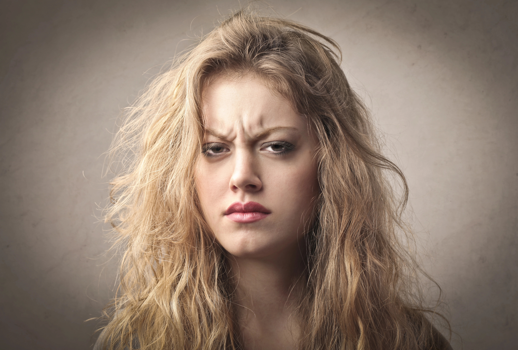 Close-up of a young woman with long, messy blonde hair, looking directly at the camera with a serious and slightly annoyed expression. She has furrowed brows and slightly pursed lips, set against a plain, neutral background.