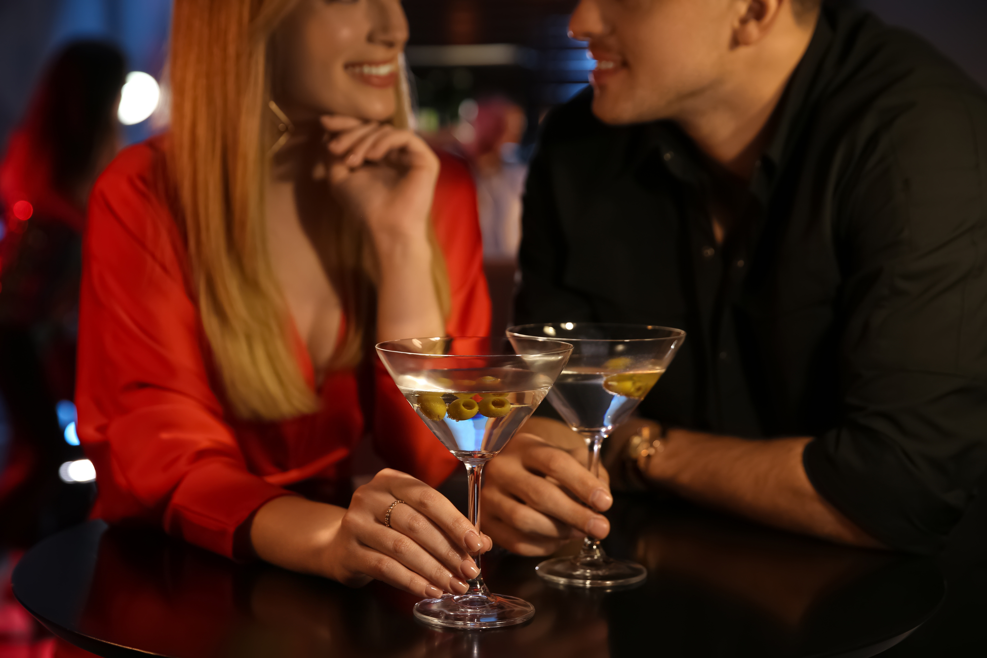A woman and a man sit at a dimly lit bar, smiling at each other. Each holds a martini garnished with olives. The woman wears a red blouse, and the man wears a black shirt. The ambiance is cozy with blurred background figures and soft lighting.