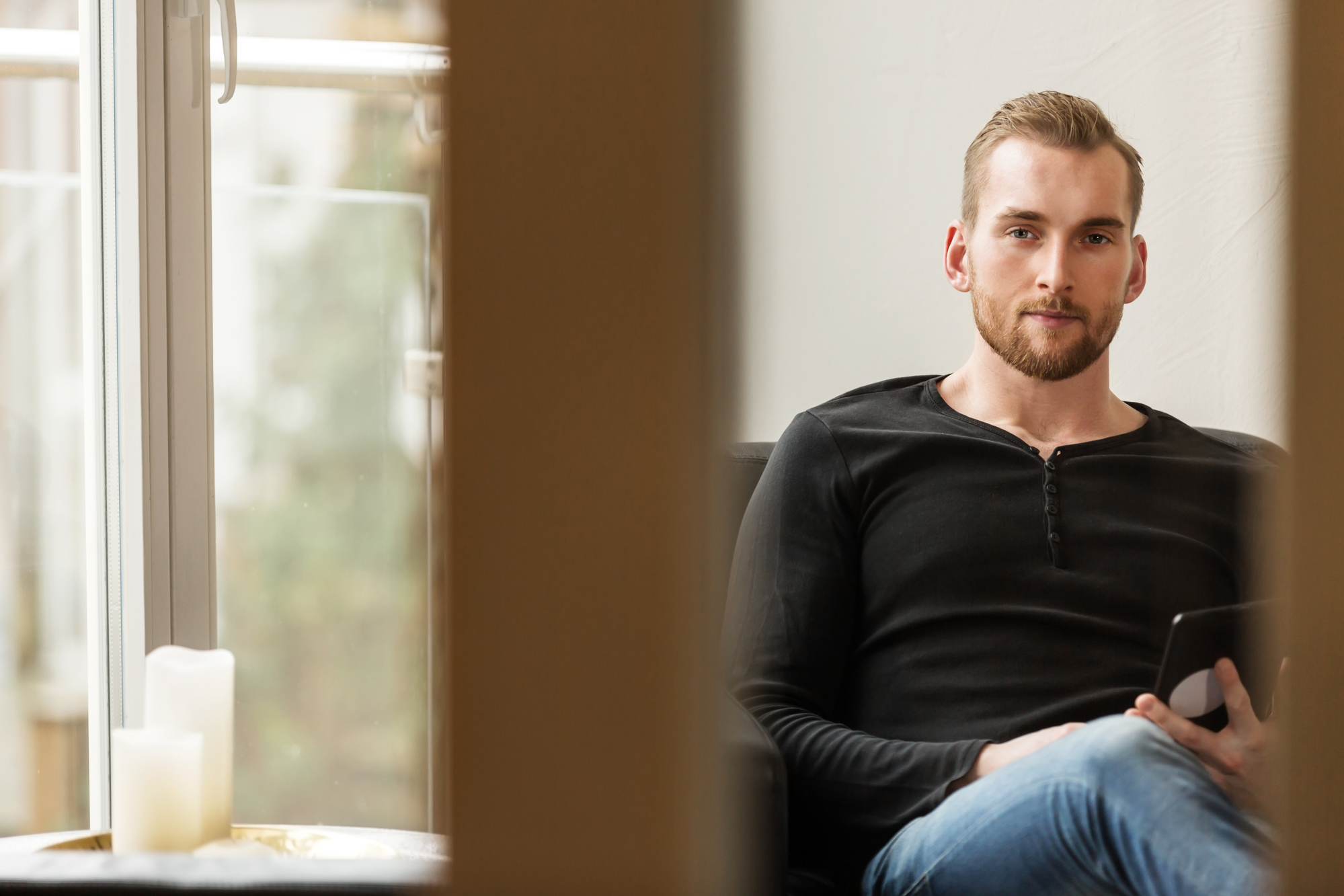 A man with short, light brown hair and a beard sits on a couch by a window holding a tablet. He is wearing a black long-sleeve shirt and jeans. Two candles are placed on the window sill next to him.
