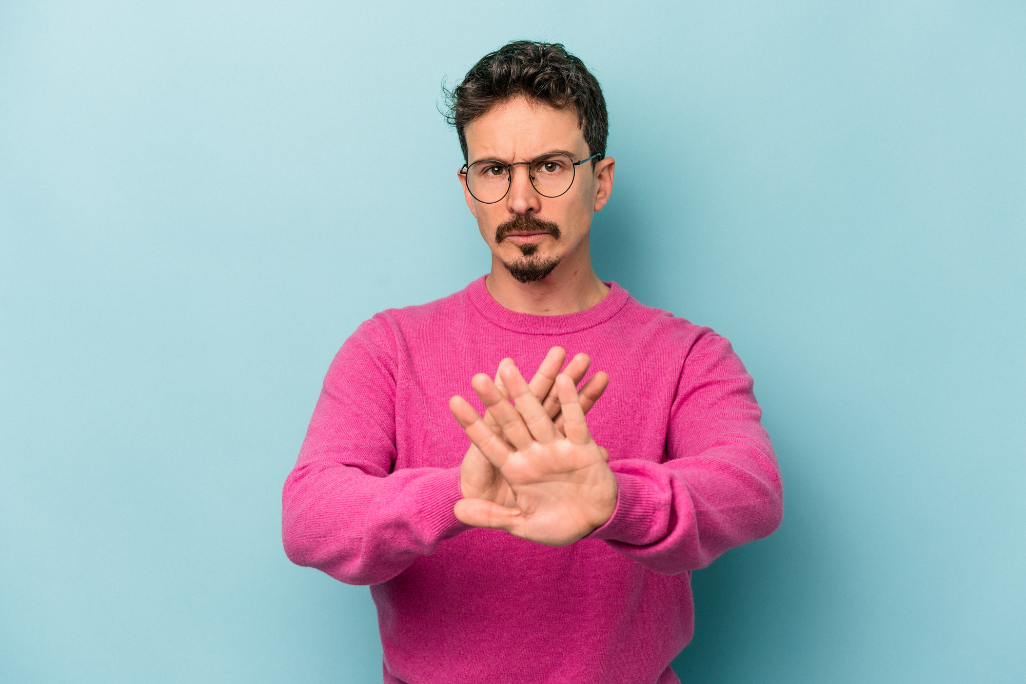 A man with short, curly hair and glasses wears a pink sweater. He stands against a blue background and has a serious expression, with his hands extended forward, palms facing out, as if signaling to stop.