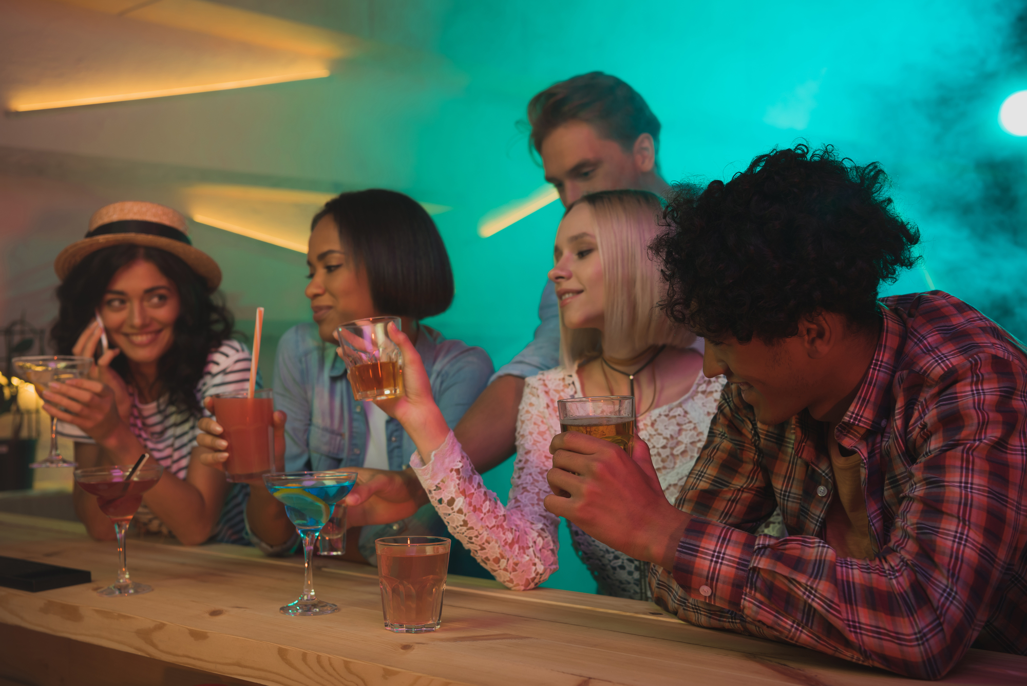 A group of five friends enjoying drinks at a bar. Two women on the left hold colorful cocktails while chatting. The other three, one man and two women, are cheerfully toasting with their drinks. The bar is dimly lit with vibrant lights in the background.