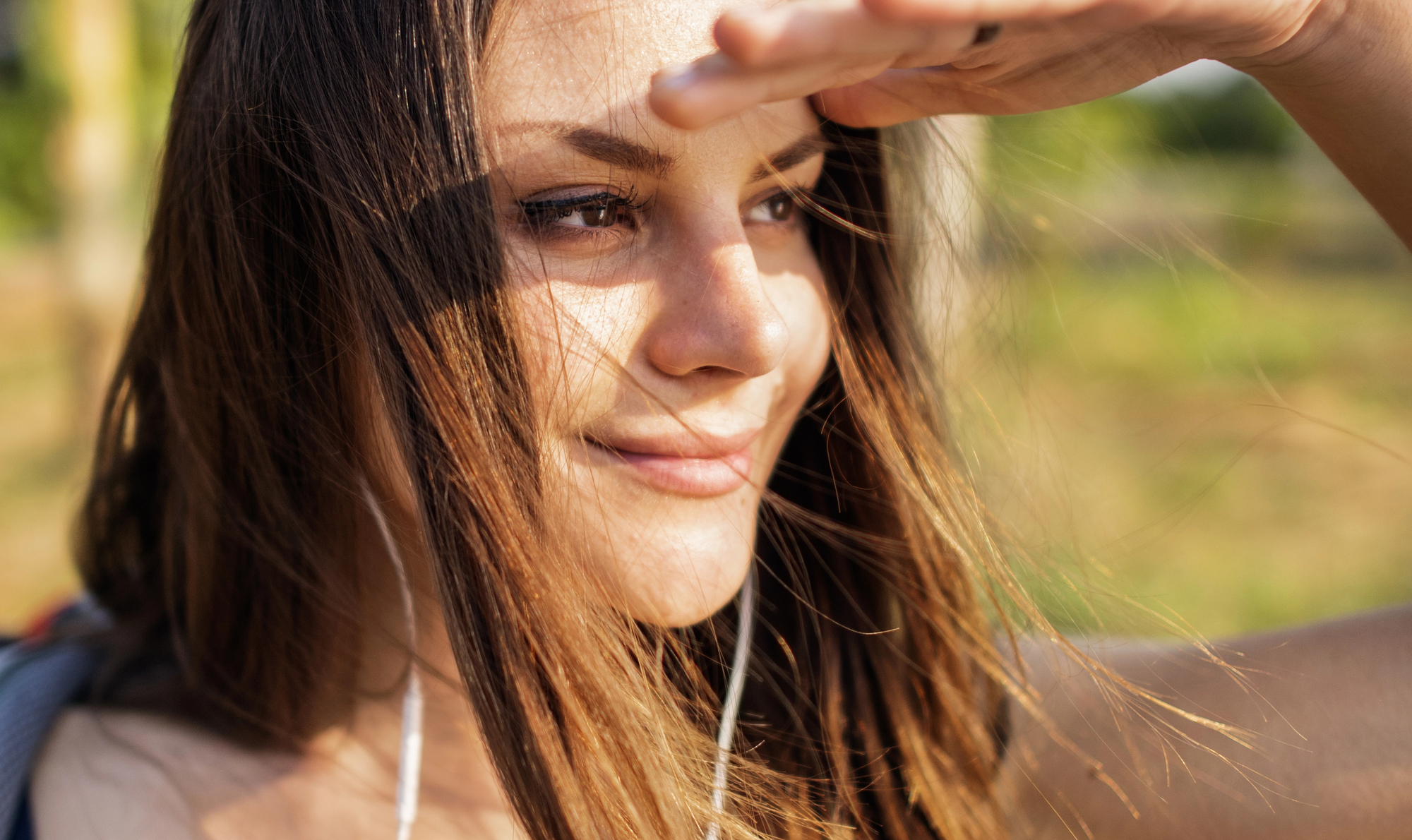 A close-up of a young woman outdoors, shielding her eyes from the sun with one hand while smiling. She has long brown hair with some strands falling across her face, and she is wearing earphones. The background is blurred, showing greenery and sunlight.