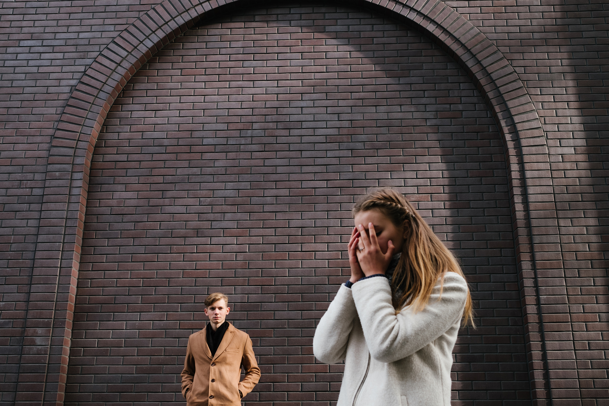 A woman in a white coat stands in the foreground, covering her face with her hands. In the background, a man in a tan coat stands with his hands in his pockets. They are in front of a large, arched brick wall. The scene appears to be emotionally charged.