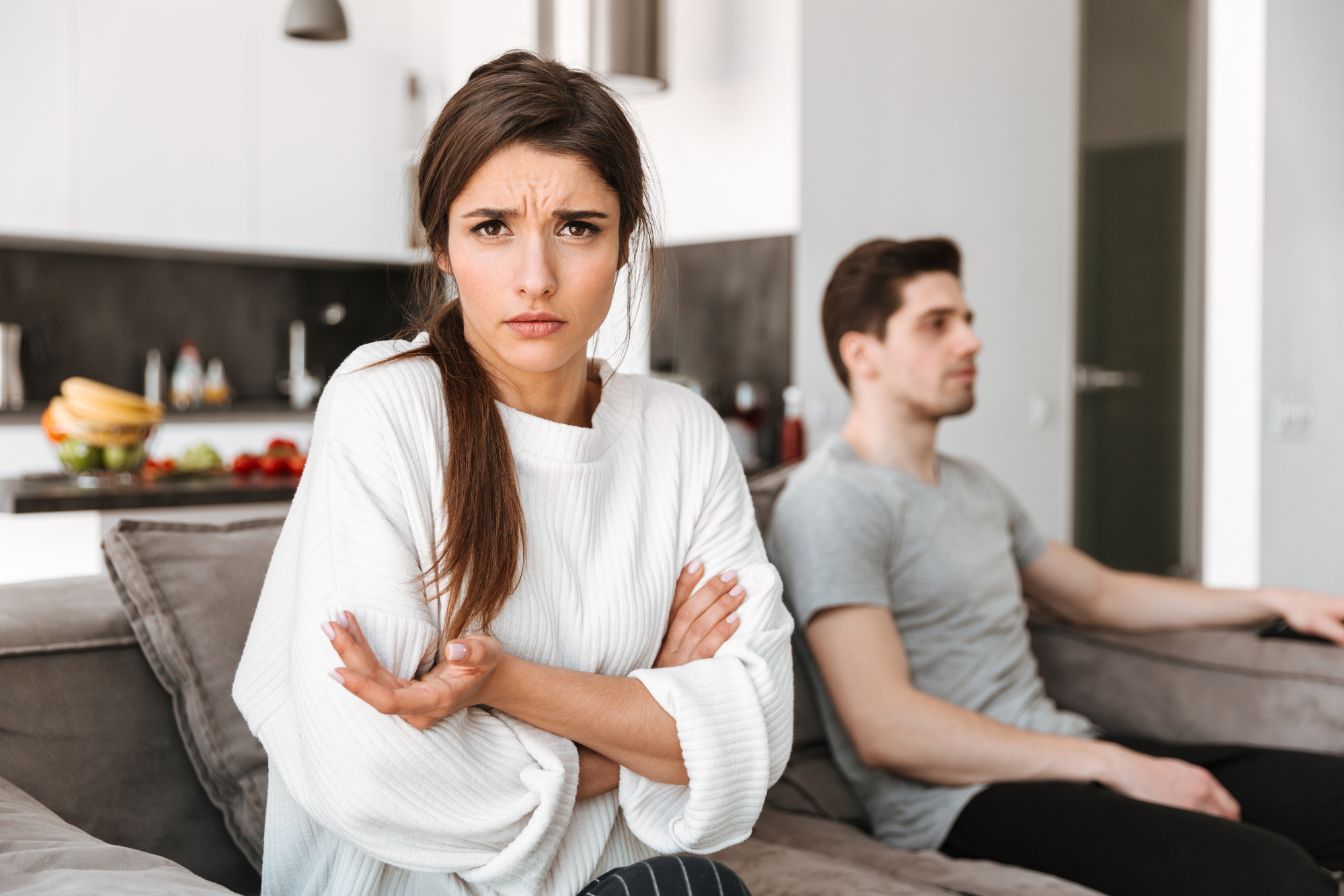 A woman with long hair in a white sweater sits on a couch with her arms crossed and an upset expression. In the background, a man in a gray t-shirt sits on the same couch looking away. They appear to be in a modern living room or kitchen setting.