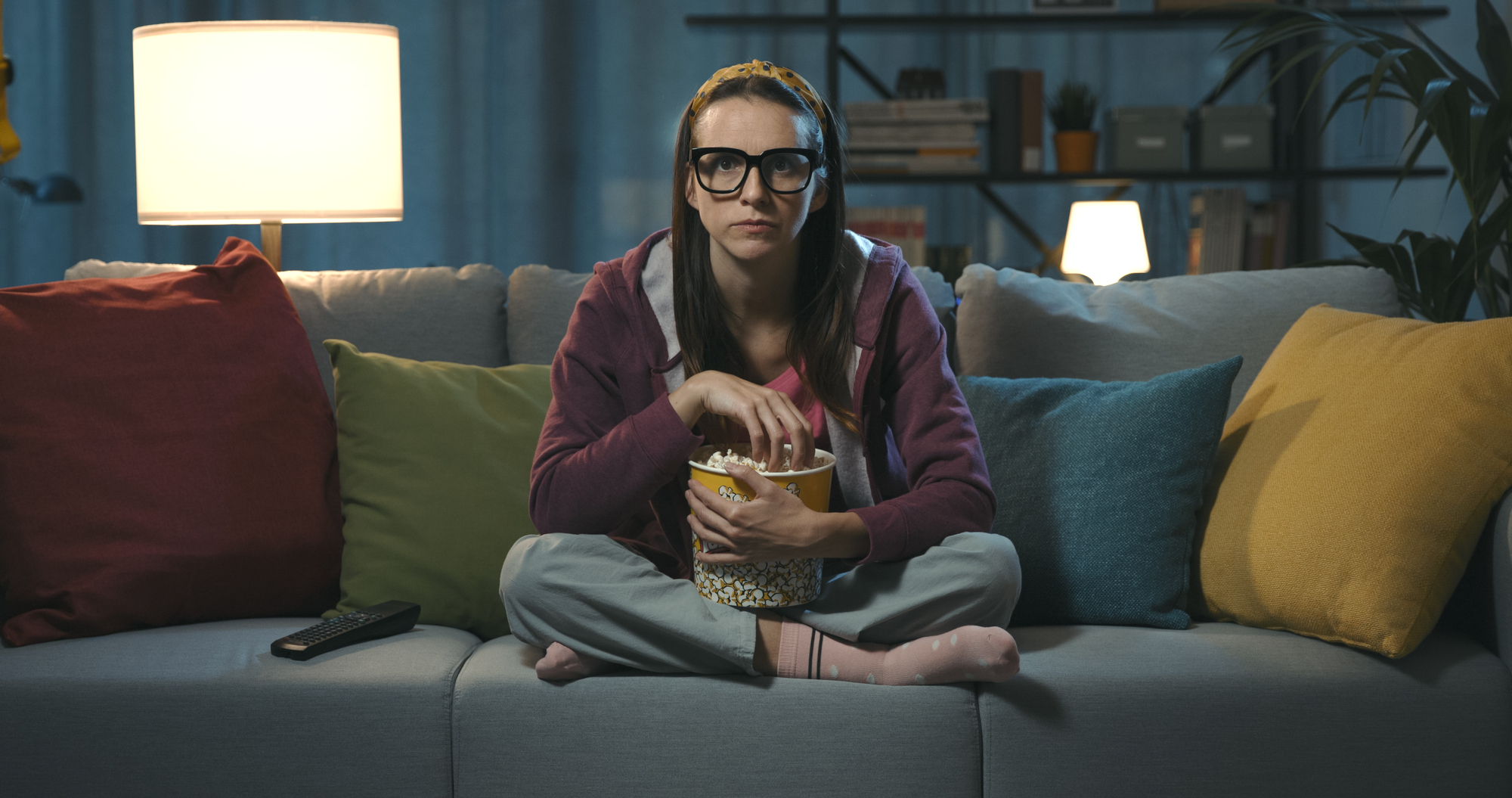 A person with glasses and a headband sits cross-legged on a couch, holding a bowl of popcorn. They are wearing a casual outfit with a hoodie and pajamas. The room is softly lit with lamps, and colorful cushions are placed on the couch. A remote control is beside them.