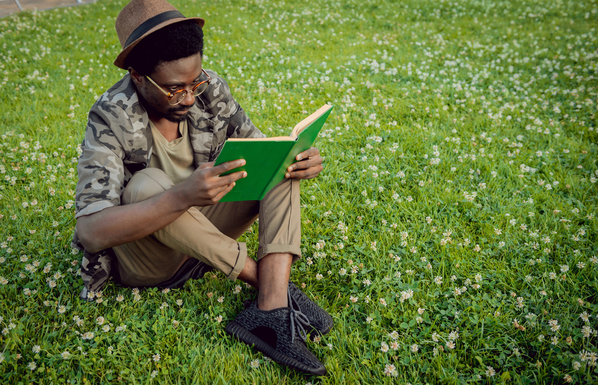 A man sits on green grass dotted with small white flowers, engrossed in reading a green book. He wears round glasses, a camouflage jacket, a brown hat, and black shoes. The relaxed outdoor setting suggests a pleasant and peaceful environment for reading.