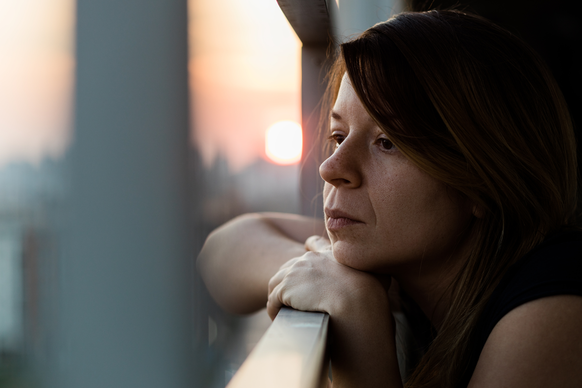 A woman with long brown hair leans on a railing, gazing pensively into the distance during sunset. The sky is tinged with warm pink and orange hues, and the soft light casts gentle shadows on her contemplative face.