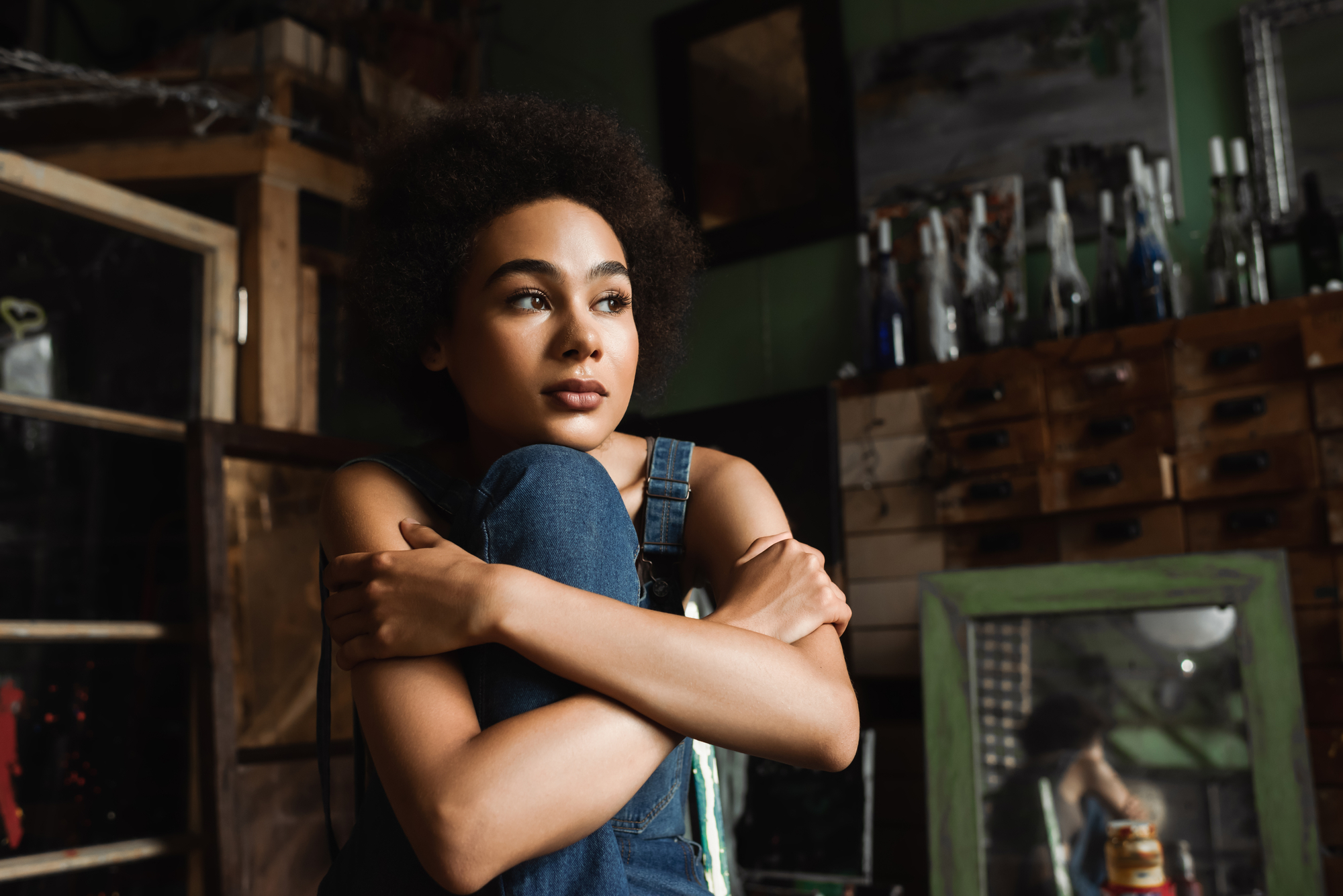 A young woman with natural curly hair sits pensively with her arms wrapped around her knees in a rustic room filled with shelves, bottles, and wooden furniture. She wears a denim outfit and looks thoughtfully into the distance.