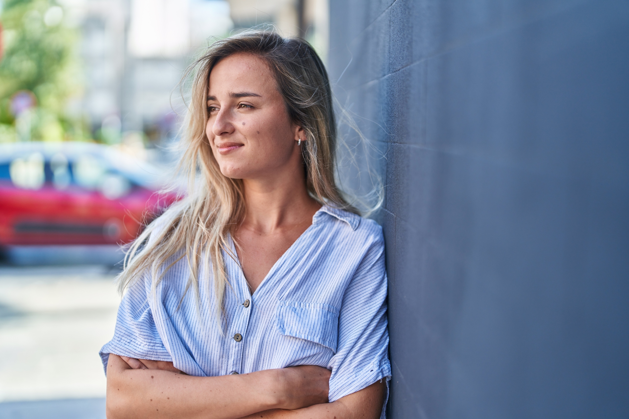 A young woman with long blonde hair and wearing a light blue, striped shirt stands against a dark gray wall. She is smiling slightly and looking to her left. A red car and blurry greenery are visible in the background.