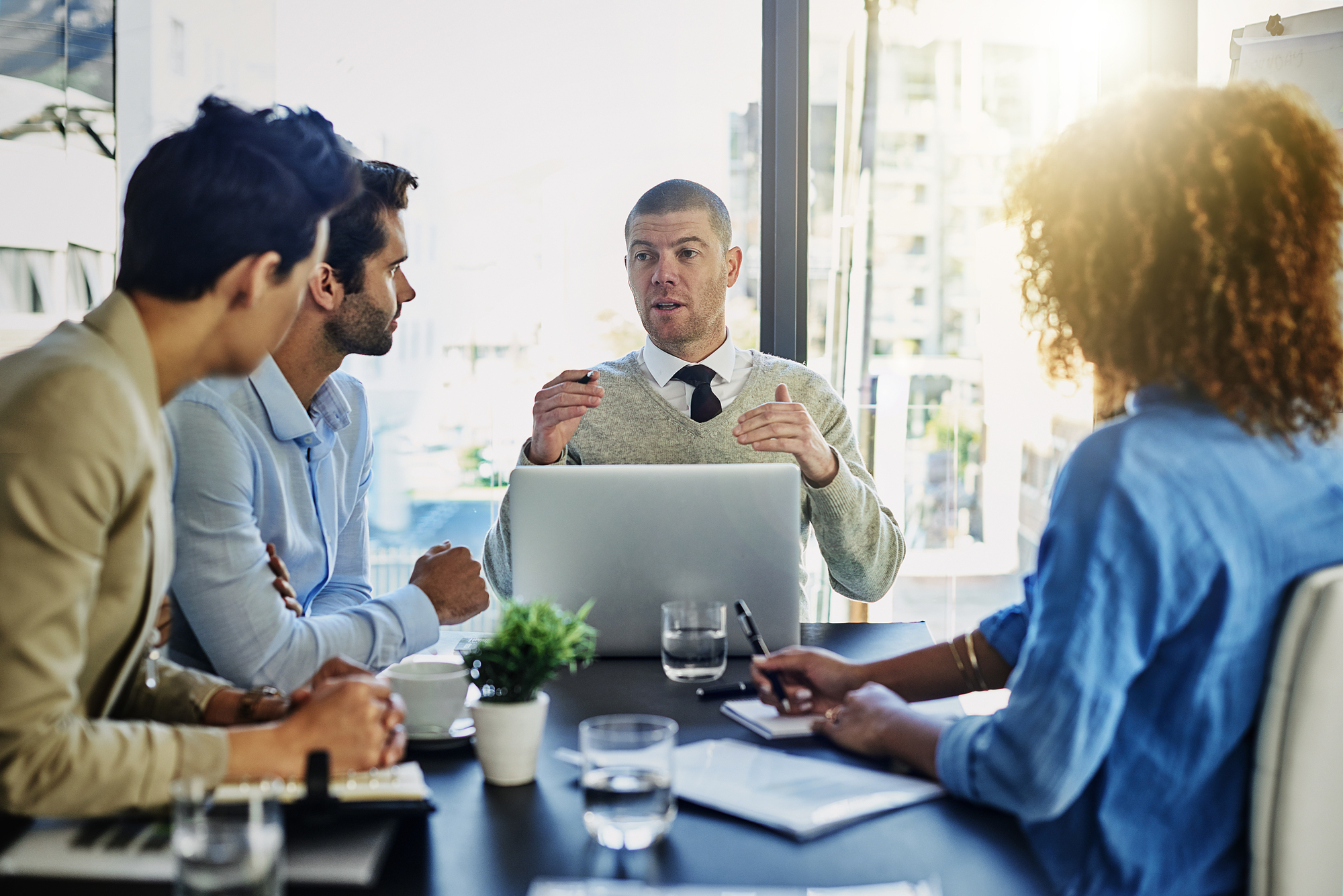 Four individuals are seated around a conference table while one person, gesturing with their hands, appears to be leading the discussion. Laptops, documents, and glasses of water are on the table. A large window in the background reveals a cityscape outside.