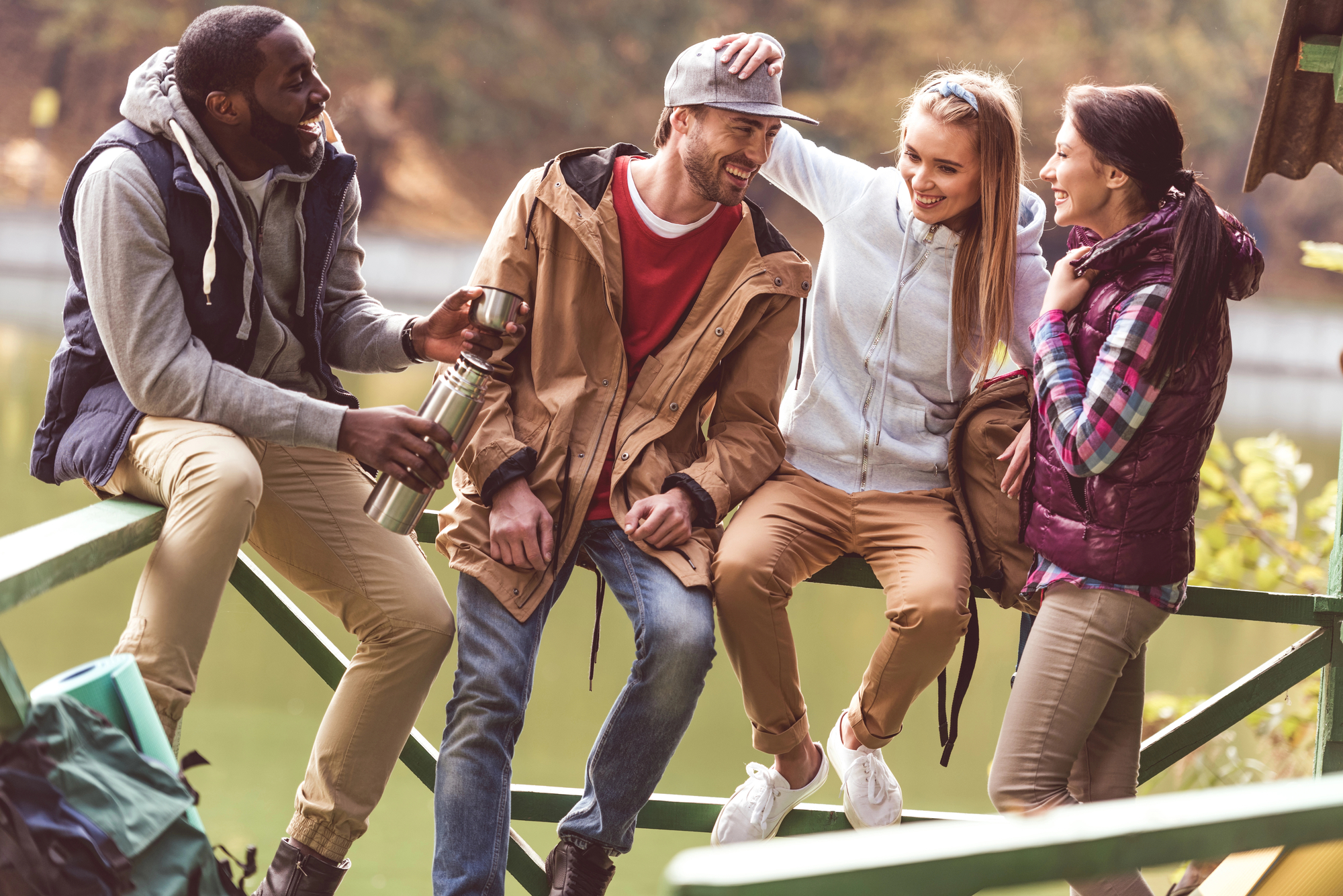 Four friends in casual outdoor clothing sit and stand around a railing near a lake, laughing and enjoying each other's company. One person is pouring a drink from a thermos, while another playfully pats a friend's head. Trees and water are visible in the background.