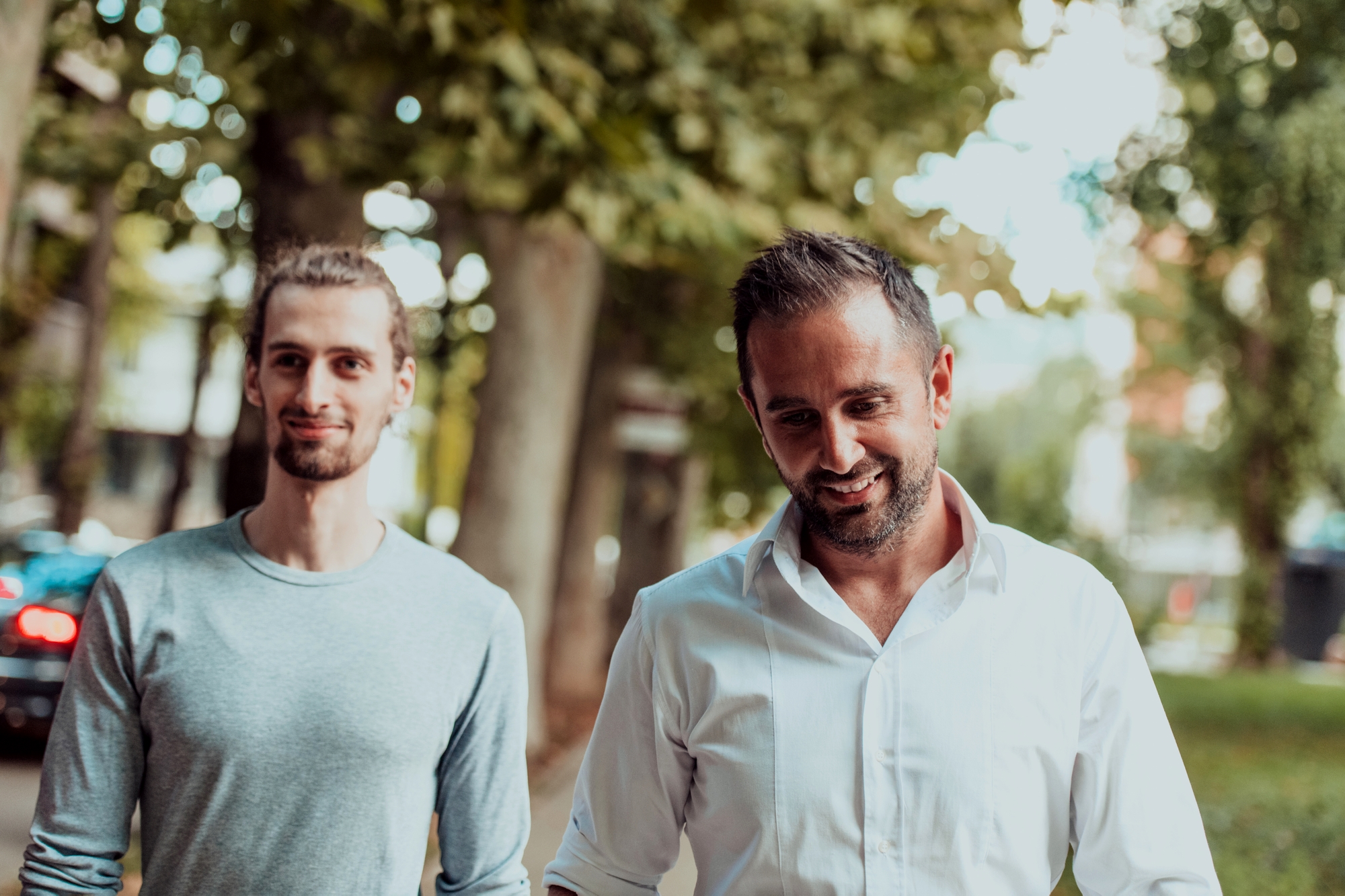 Two men walking outdoors on a tree-lined path. The man on the right is wearing a white button-down shirt and smiling, while the man on the left is wearing a gray long-sleeved shirt and also smiling. They appear to be engaged in conversation, with blurred nature in the background.