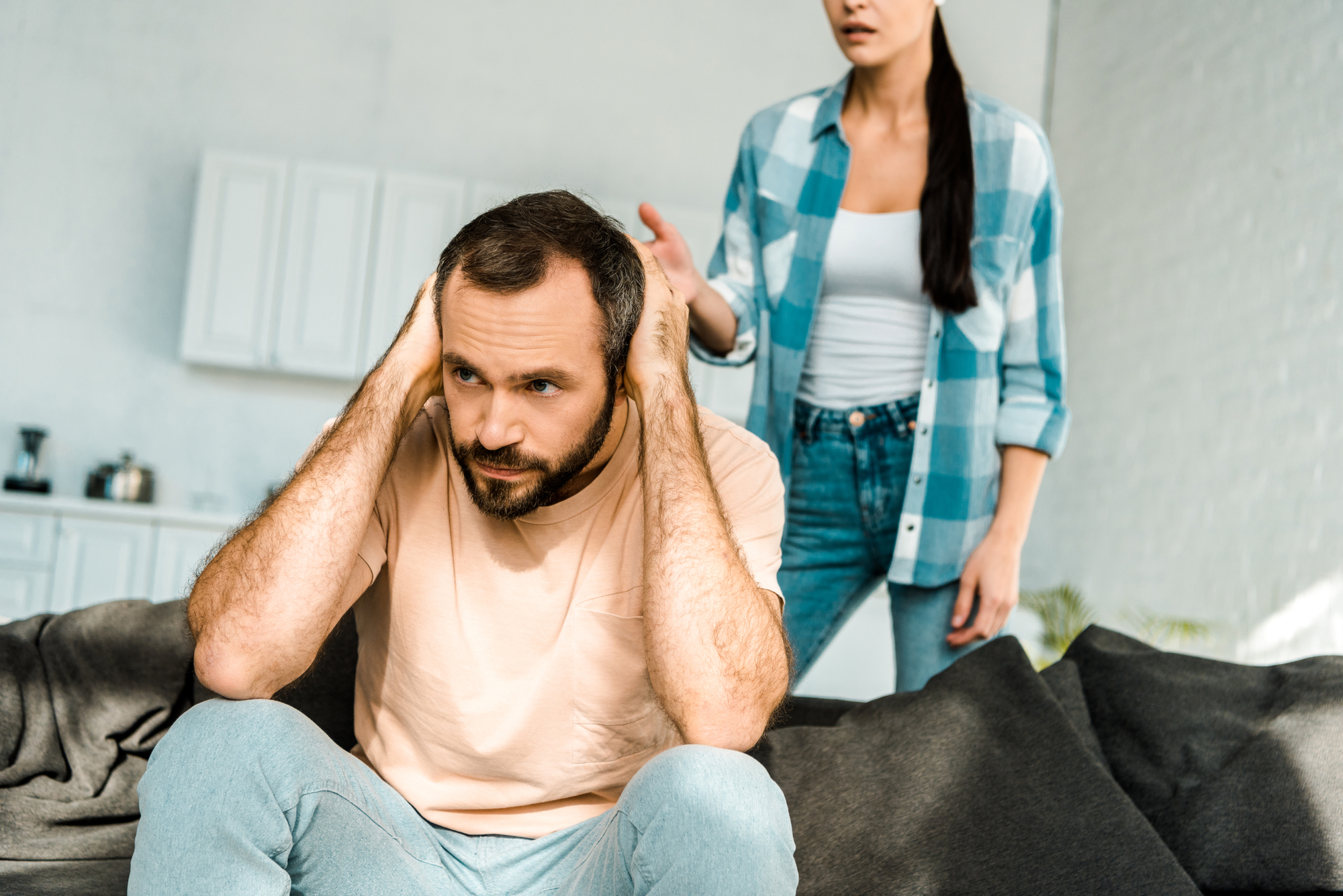 A man sits on a couch, covering his ears and looking distressed. A woman stands behind him with her hand raised, appearing to speak or argue. They are in a modern, bright living room with a kitchen in the background.