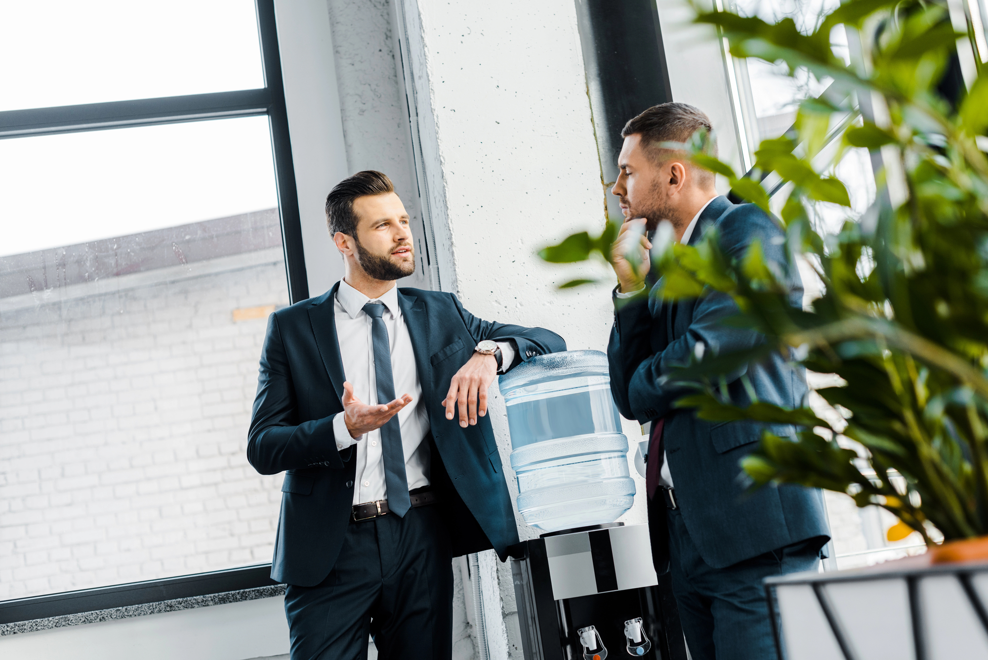 Two men in business suits are talking near a water cooler in an office setting. One man gestures with his hand while the other listens, touching his chin. The office has large windows and a plant in the foreground.