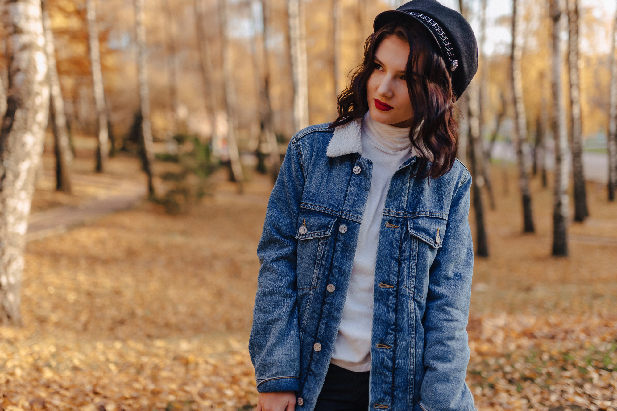A young woman stands in a sunlit park with autumn trees, wearing a denim jacket, white turtleneck, black hat, and red lipstick. She looks to her left with a serene expression, surrounded by fallen leaves on the ground.