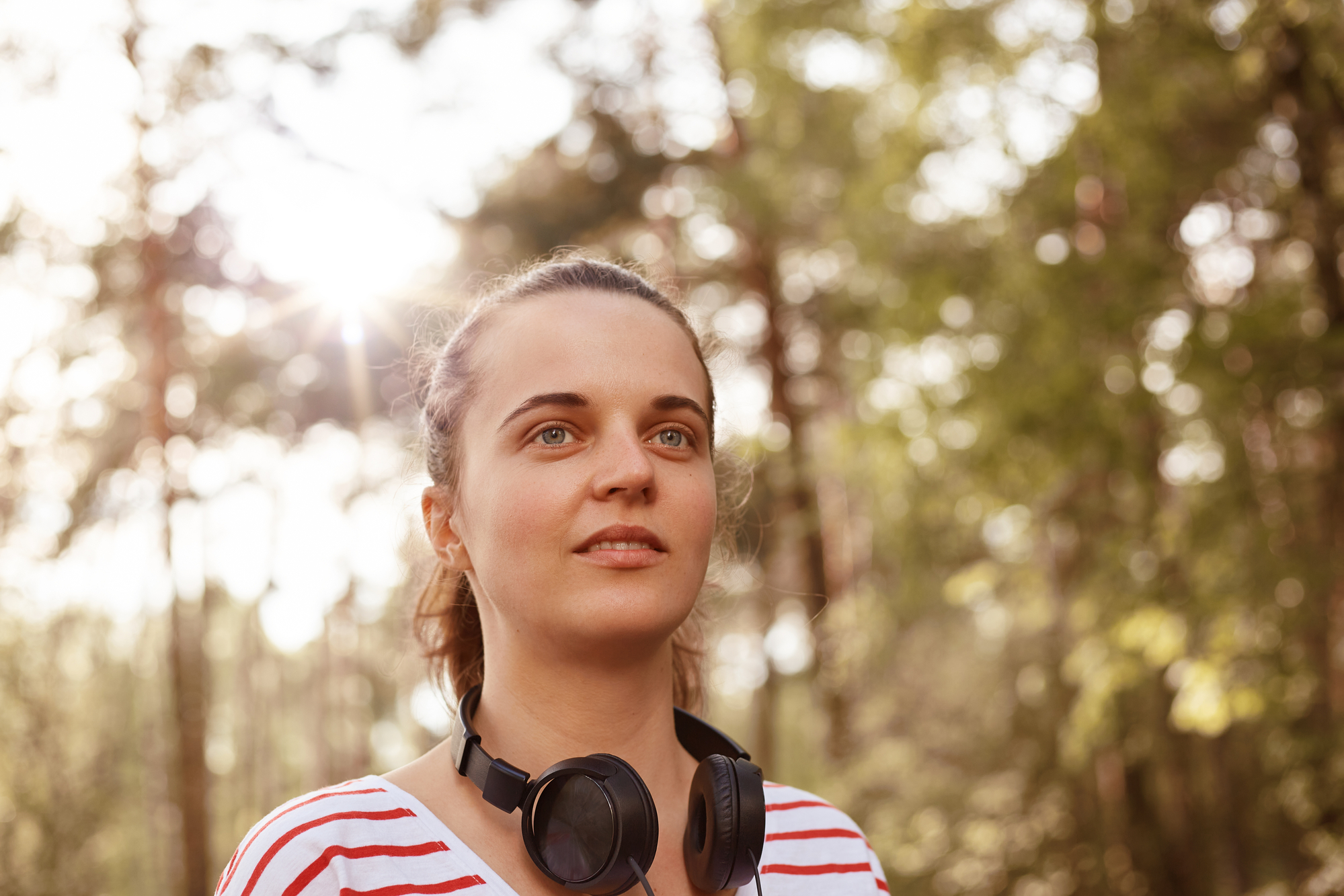 A person with a thoughtful expression stands outdoors in a sunlit forest wearing a red and white striped shirt and black headphones around their neck. Trees and sunlight filtering through the leaves create a serene background.