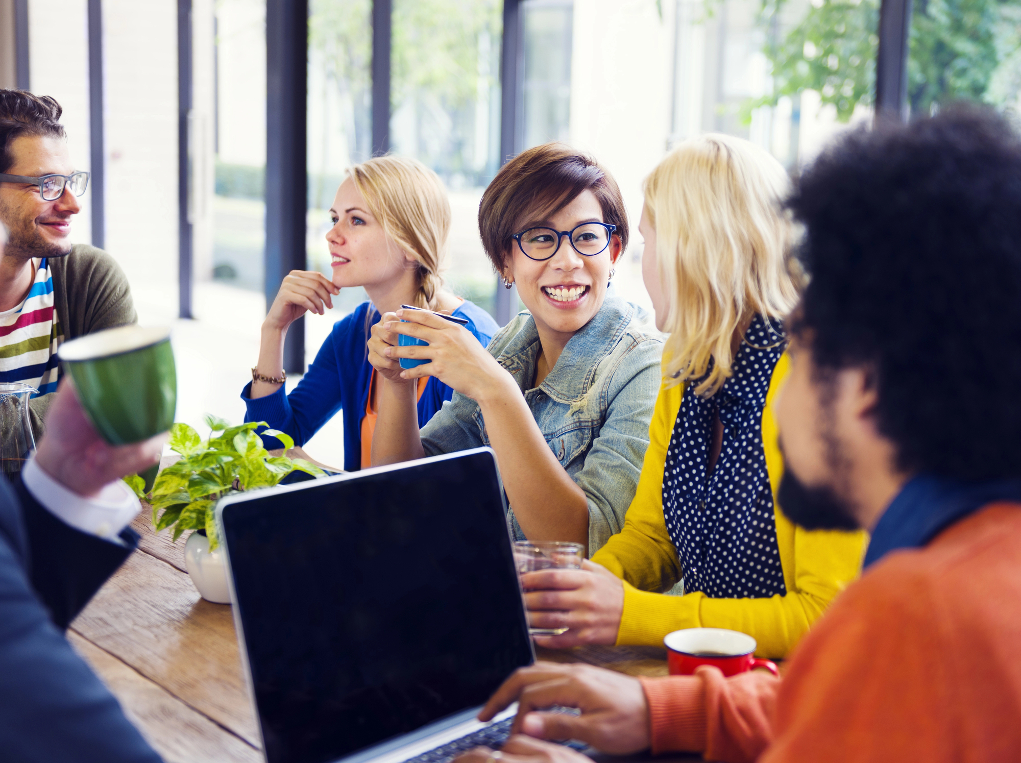 A diverse group of people, sitting around a table, engaged in a lively conversation. They are all smiling and appear relaxed. One person holds a laptop, while others have coffee mugs. There's a small plant on the table, and large windows in the background.