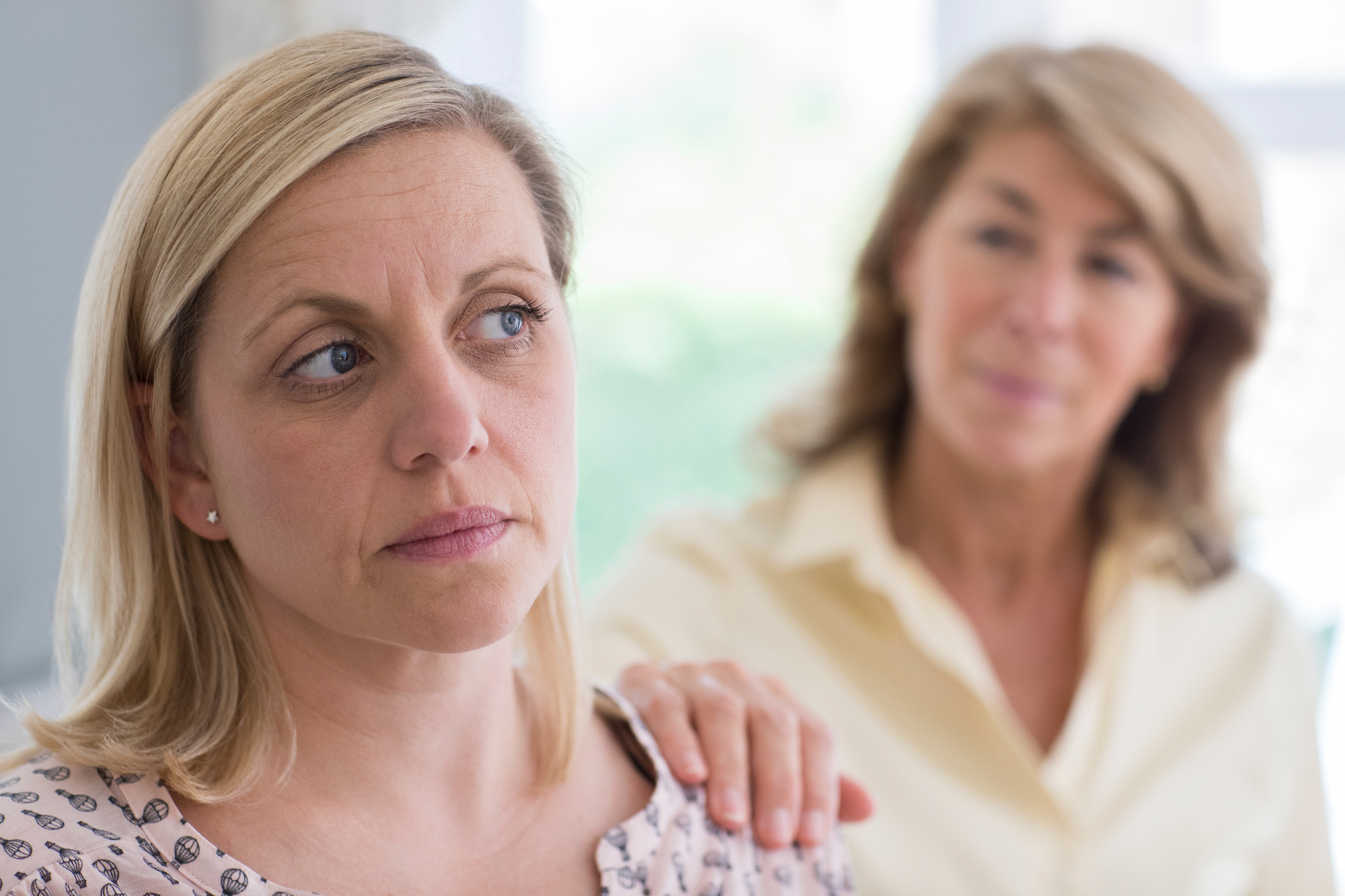 A woman with a somber expression looks away while another older woman gently places her hand on her shoulder in a comforting gesture. The two are indoors, with a blurred background suggesting a bright room.