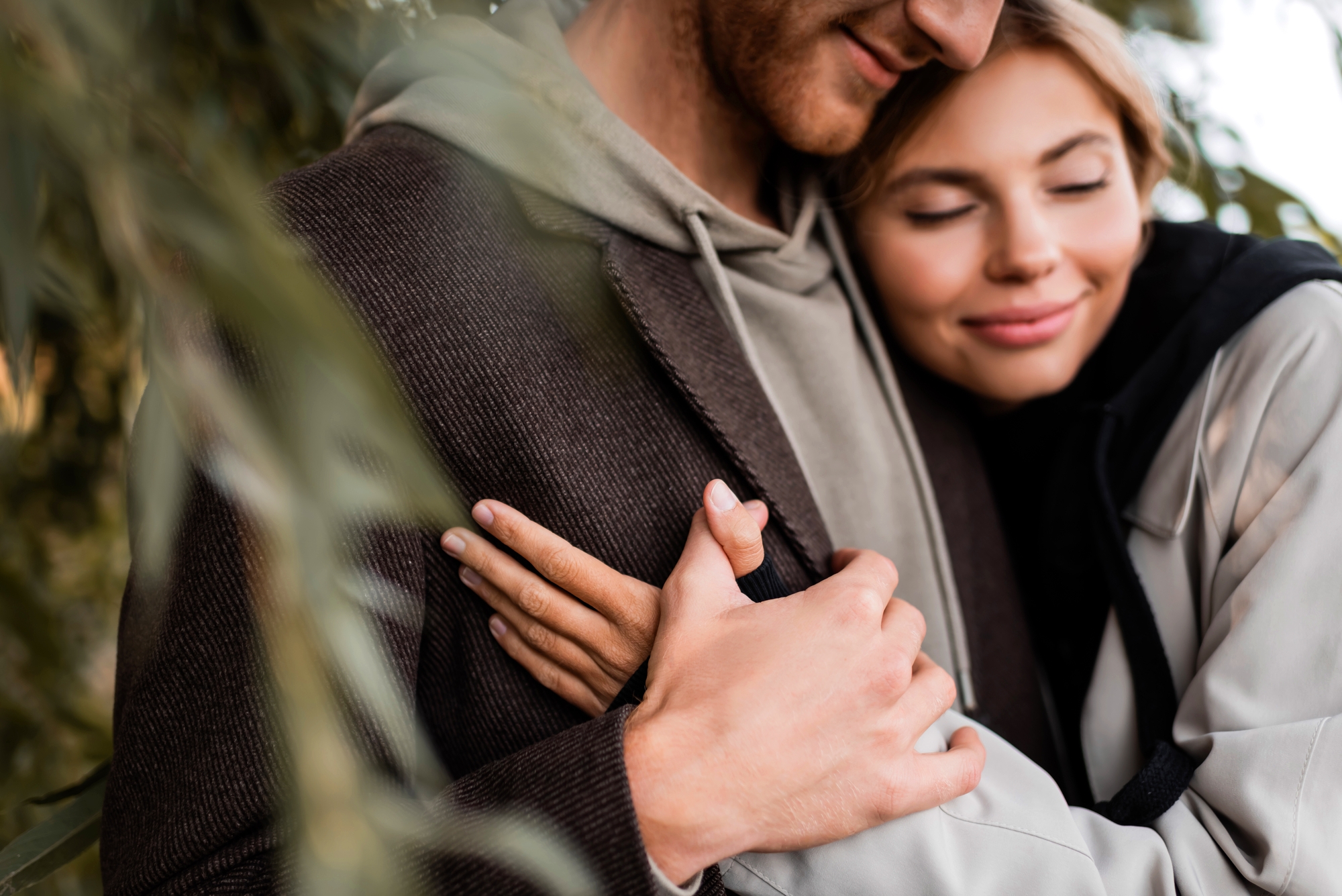 A close-up of a couple embracing. The woman, with a content smile, has her eyes closed and is hugging the man tightly. The man's face is partially visible, and he is wearing a gray hoodie and a brown jacket. Both seem to be outdoors, surrounded by greenery.
