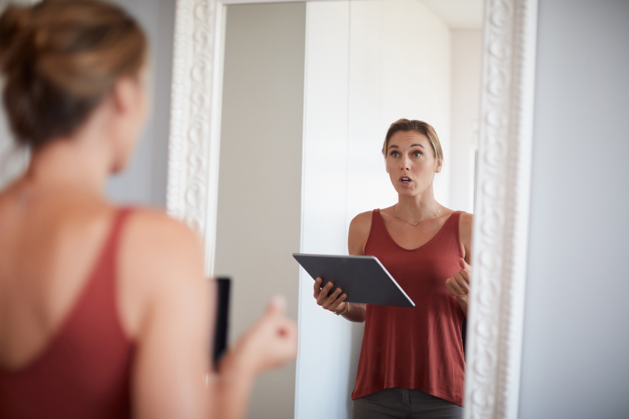 A woman stands in front of a mirror, holding a tablet and speaking. She is wearing a sleeveless red top, with her hair tied back. Her expression suggests she is practicing a speech or presentation. The background is a softly lit, neutral-toned room.