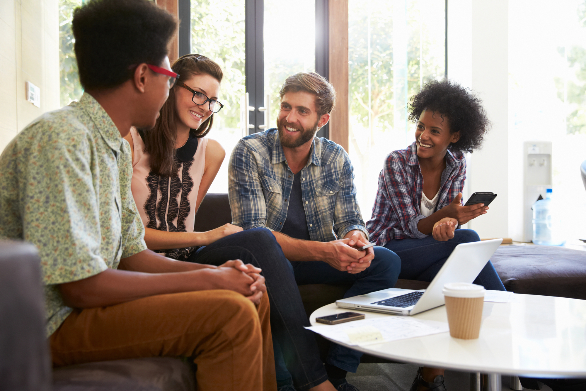 Four diverse individuals are seated on a couch in a relaxed environment, engaging in a friendly conversation. One person holds a smartphone, and an open laptop is on the table in front of them, alongside a paper cup and some documents. Natural light fills the room.